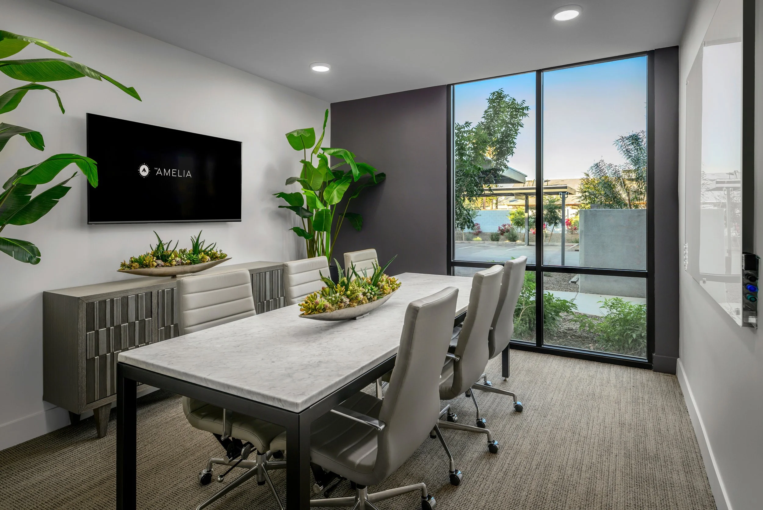 Modern conference room with a marble table, six white chairs, potted plants, a large window, and a wall-mounted TV displaying 'Amelia' logo.