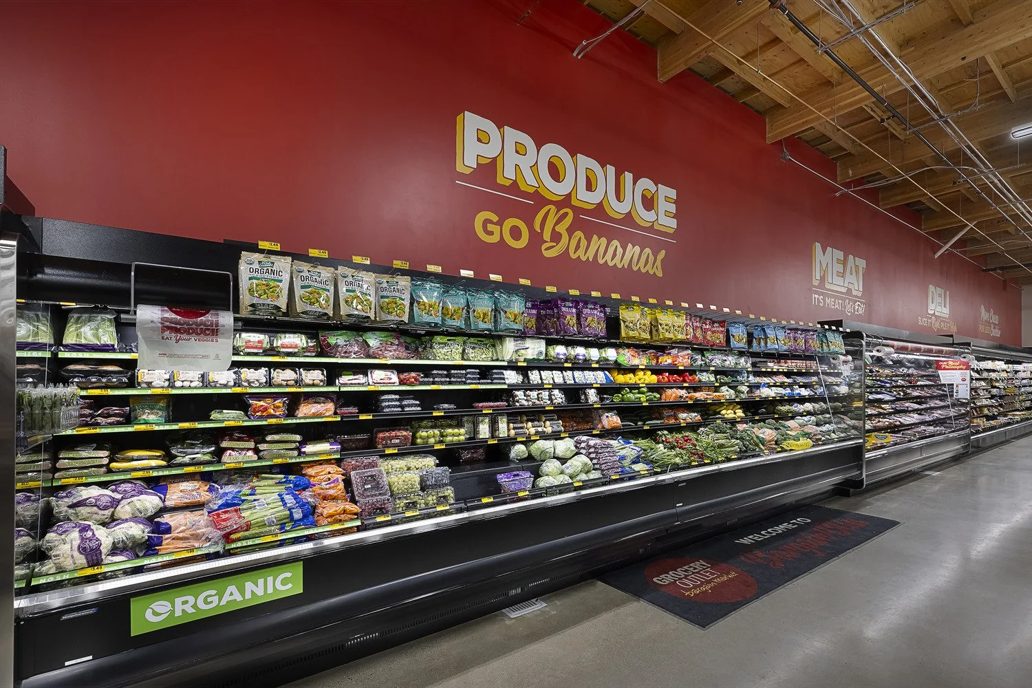 Grocery store produce aisle with vegetables and fruits on shelves, and signs on the wall reading "Produce Go Bananas" and "Meat", in a modern store with a concrete floor and wooden ceiling.