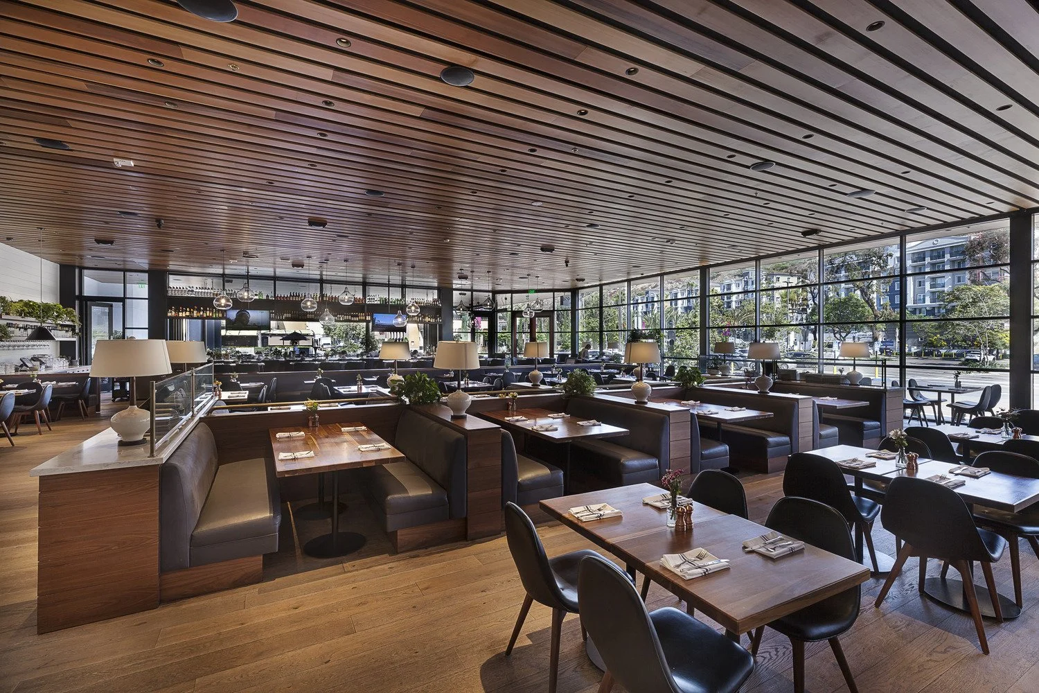 Interior of a modern restaurant with wooden floors, a wooden ceiling, large glass windows, and mixed seating including tables with black chairs and booths with dark leather seats, decorated with lamps and small flower arrangements.
