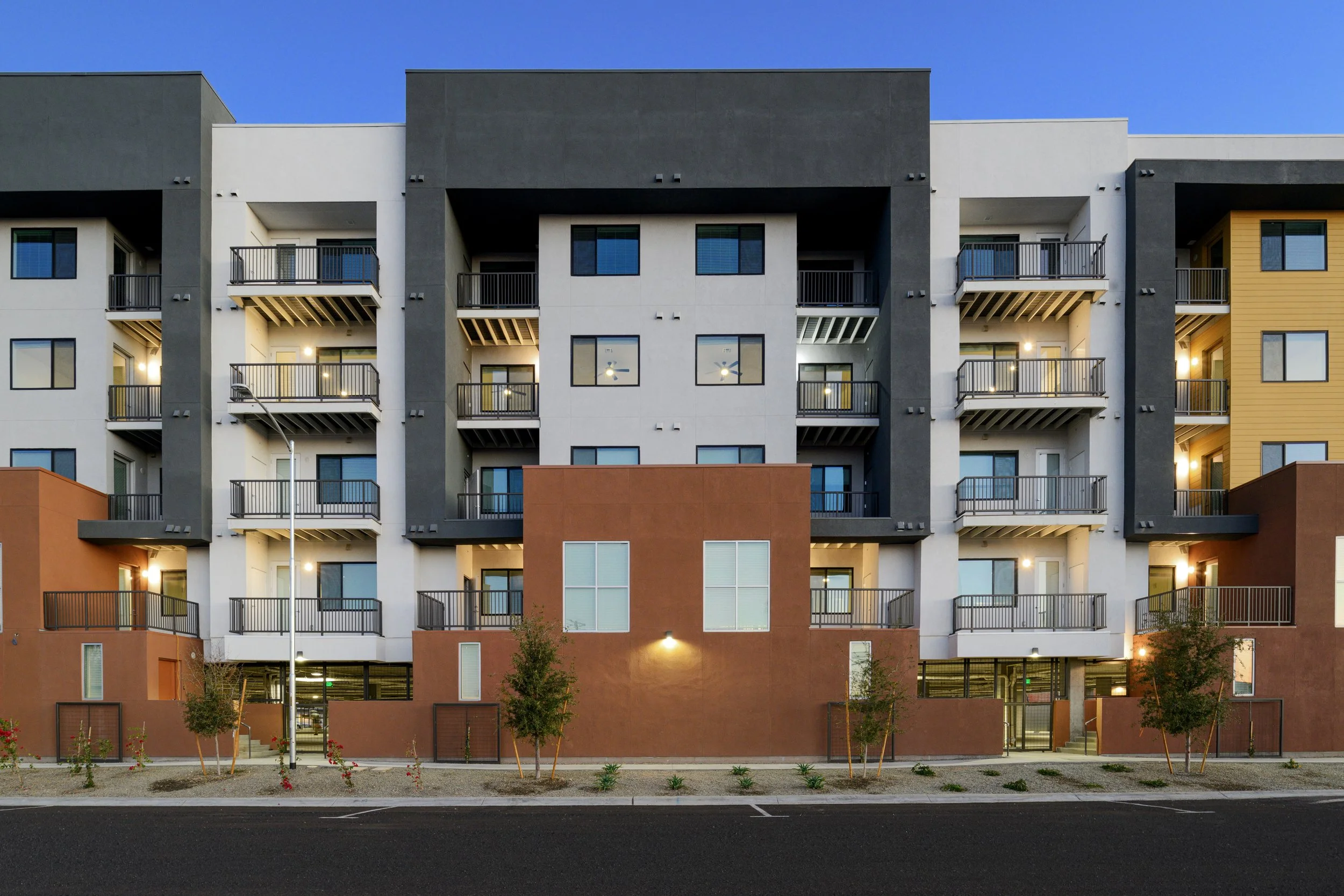 Modern multi-story apartment building with balconies, illuminated windows, and landscaped front yard during dusk.