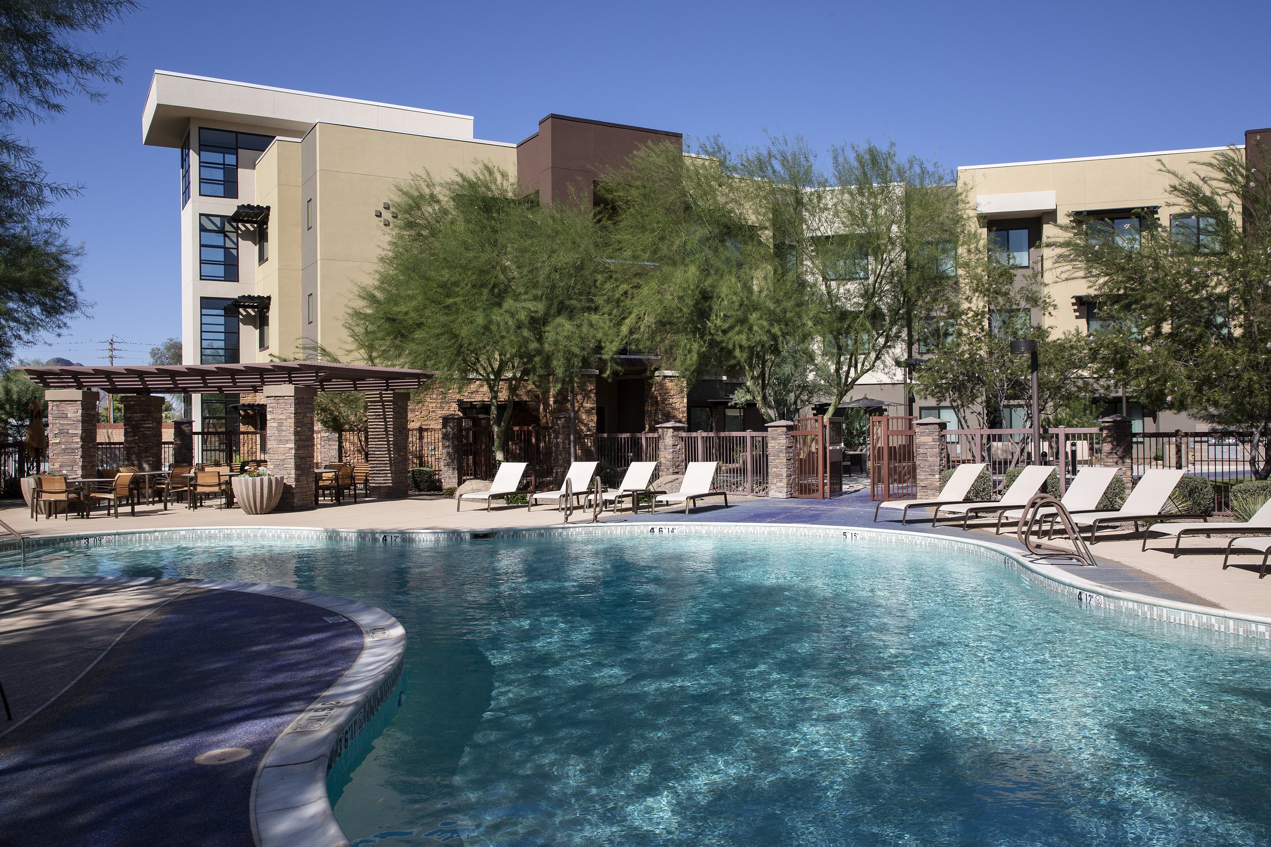 Empty swimming pool area with lounge chairs, a shaded seating area, and modern apartment buildings in the background under a clear blue sky.