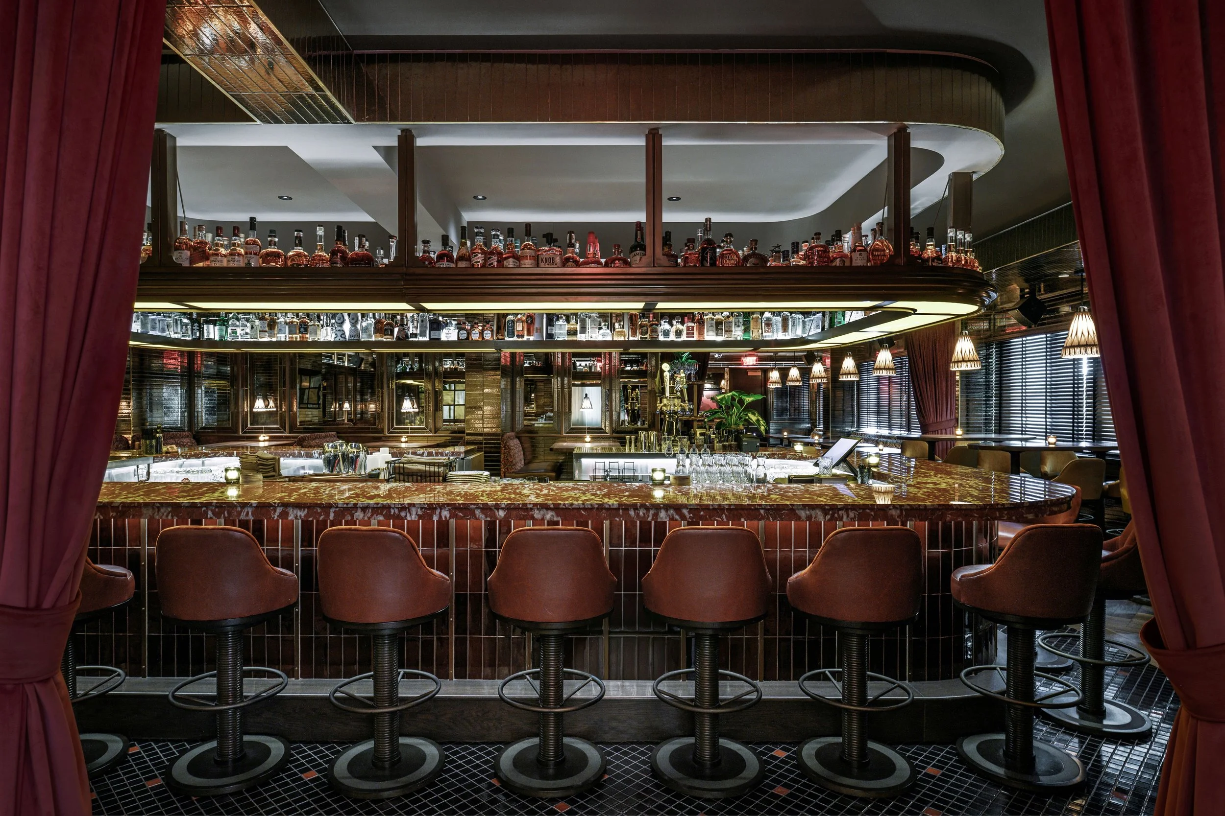 Inside a luxurious bar with a marble countertop, six brown bar stools, shelves stocked with liquor bottles above, warm lighting, and large windows with blinds.