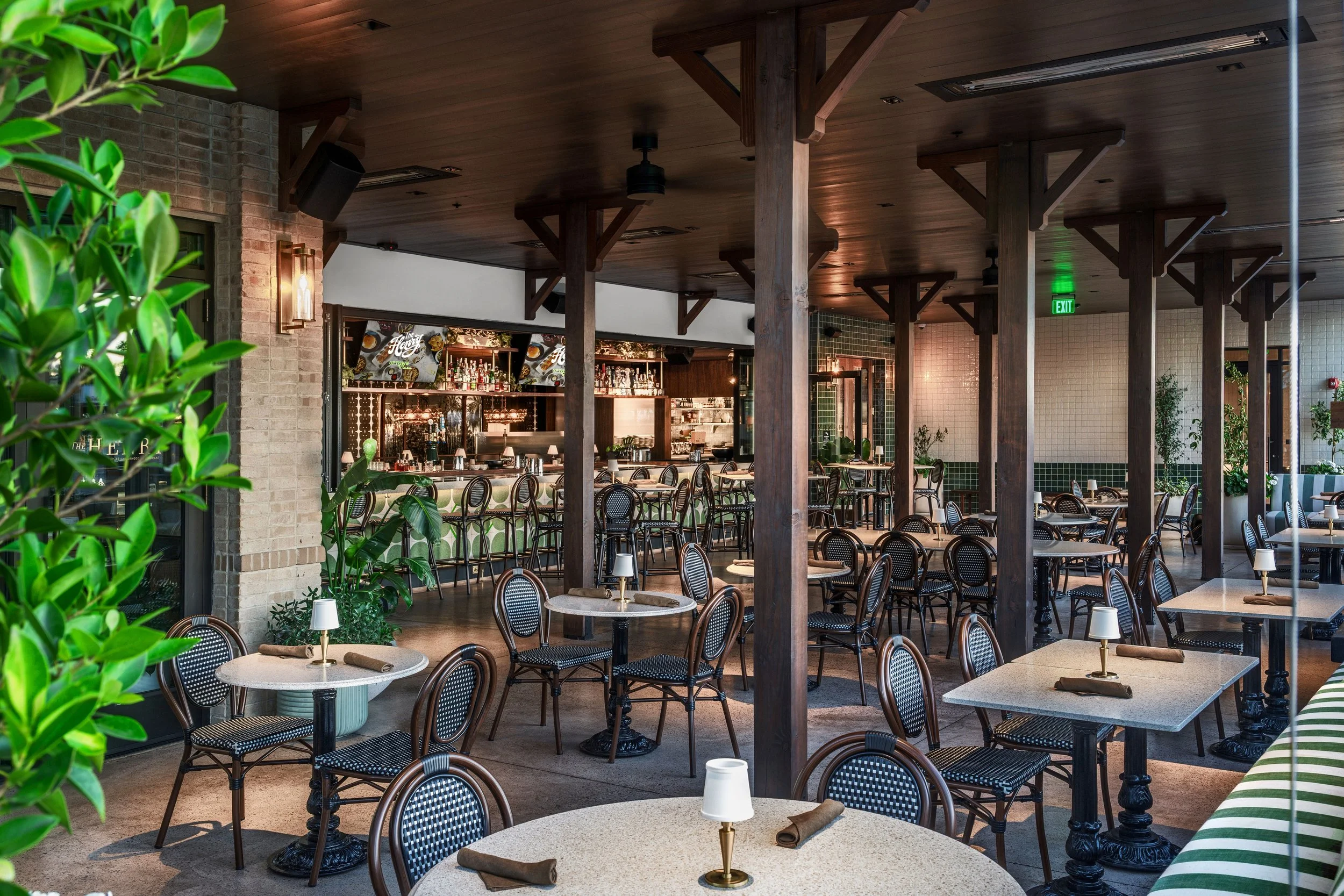Interior of a restaurant with round tables, chairs with black woven seats, a bar area in the back, and green plants decorating the space.