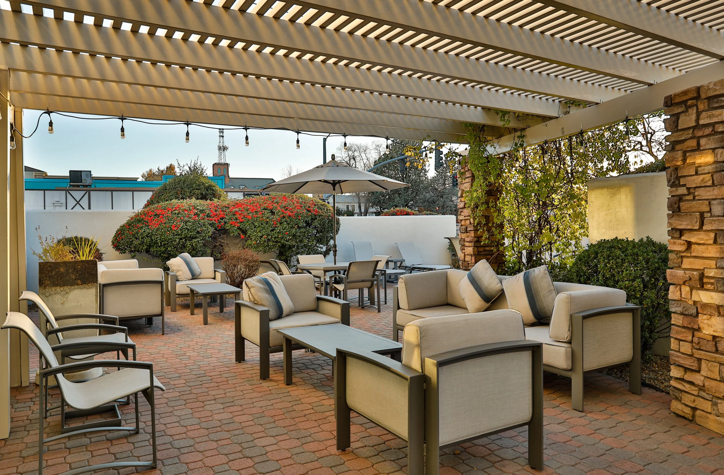 Outdoor patio area with seating, couches, armchairs, tables, and umbrellas, surrounded by plants and brick paving.