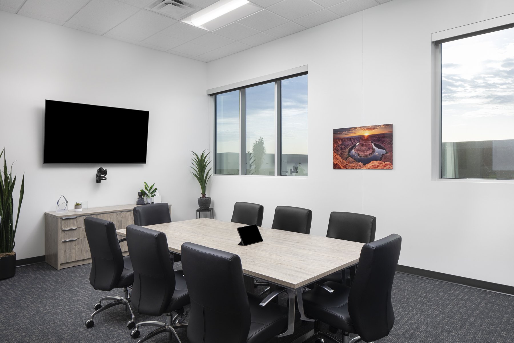 Modern conference room with a large wooden table surrounded by black leather chairs, a TV screen on the wall, windows letting in natural light, and a picture of a canyon landscape with a river at sunset.