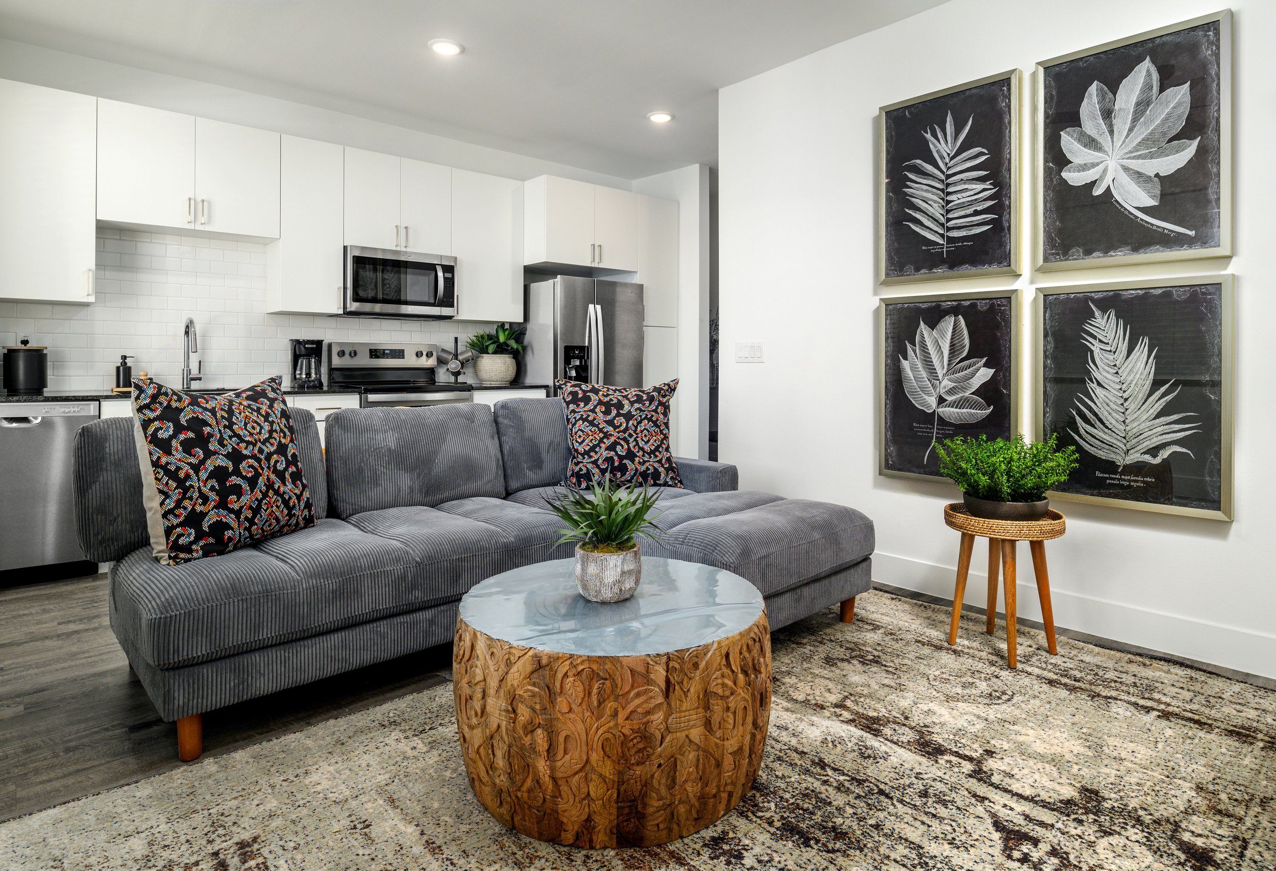 Living room with gray sofa, patterned pillows, wooden and marble coffee table, green plants, and wall art of four blackboards with white leaf illustrations, open kitchen with white cabinets and stainless steel appliances.