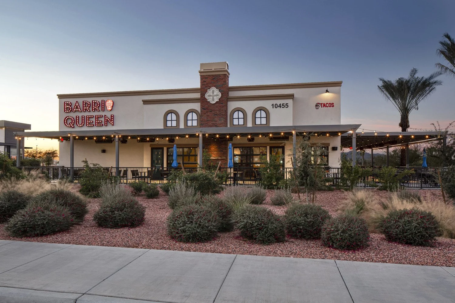 Exterior of Barrio Queen restaurant with a sign illuminated in red lights, outdoor seating area with string lights and umbrellas, desert landscaping with bushes and grasses, at sunset.