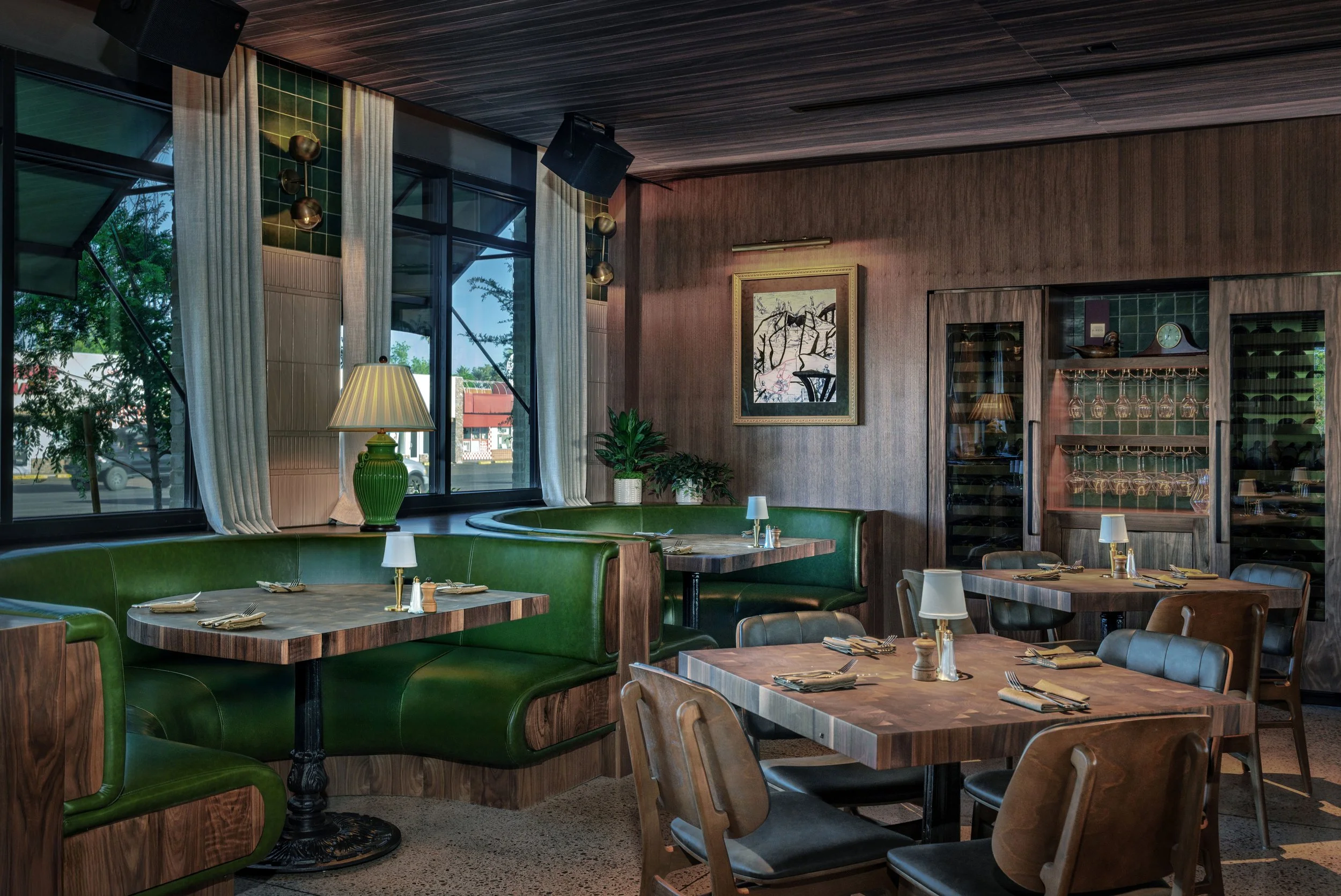 Interior of a restaurant with green booth seating, wooden tables, and table settings including napkins, silverware, and small lamps. Large windows with white curtains, a wooden wall with framed art, and a glass cabinet with wine glasses are visible.