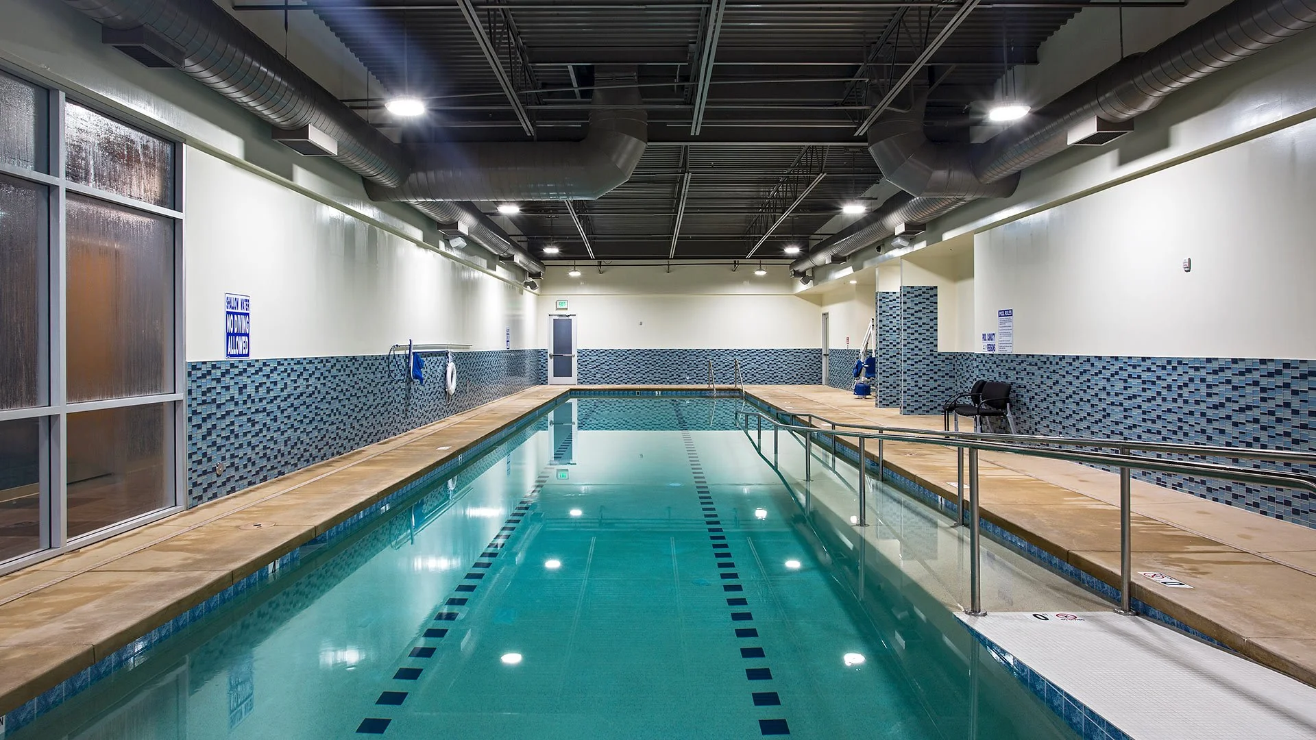 Indoor swimming pool with lane markers, safety railings, and a tiled corner, illuminated overhead, with frosted window on the left and a door at the end.