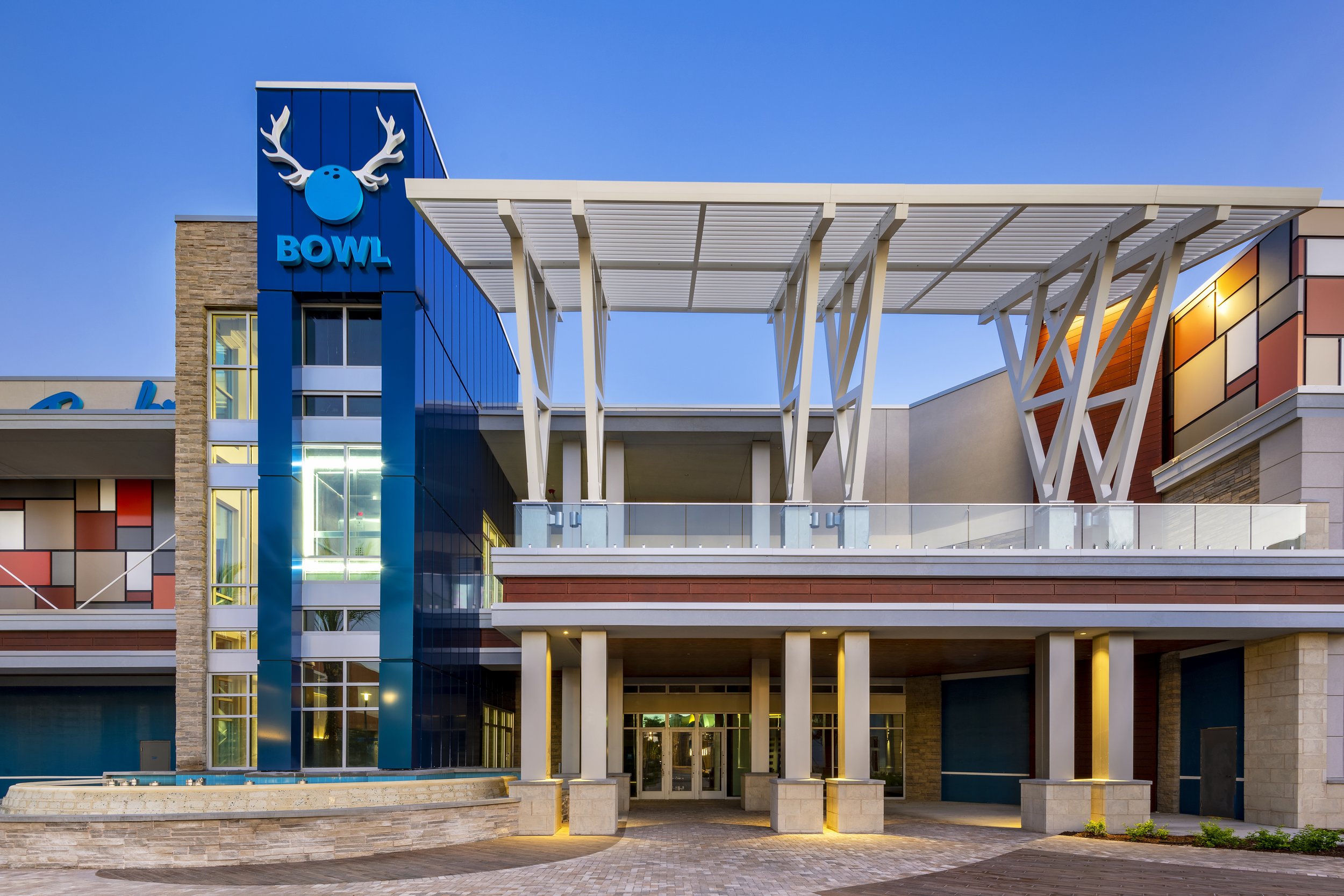 Modern shopping mall with architectural design, blue and orange panels, glass railings, and a logo with a blue face with white antlers and the word 'Bowlero' on the left side.