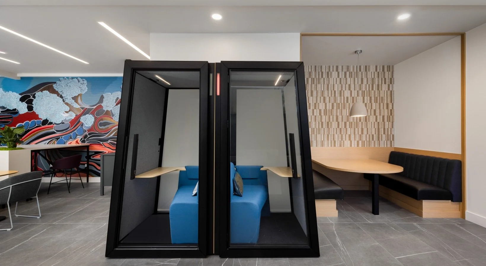 Modern office or lounge with two black private work booths with blue sofas inside, a booth with a grey interior, a mural wall with colorful abstract art, and a corner nook with a wooden table, black bench seating, and a pendant light.