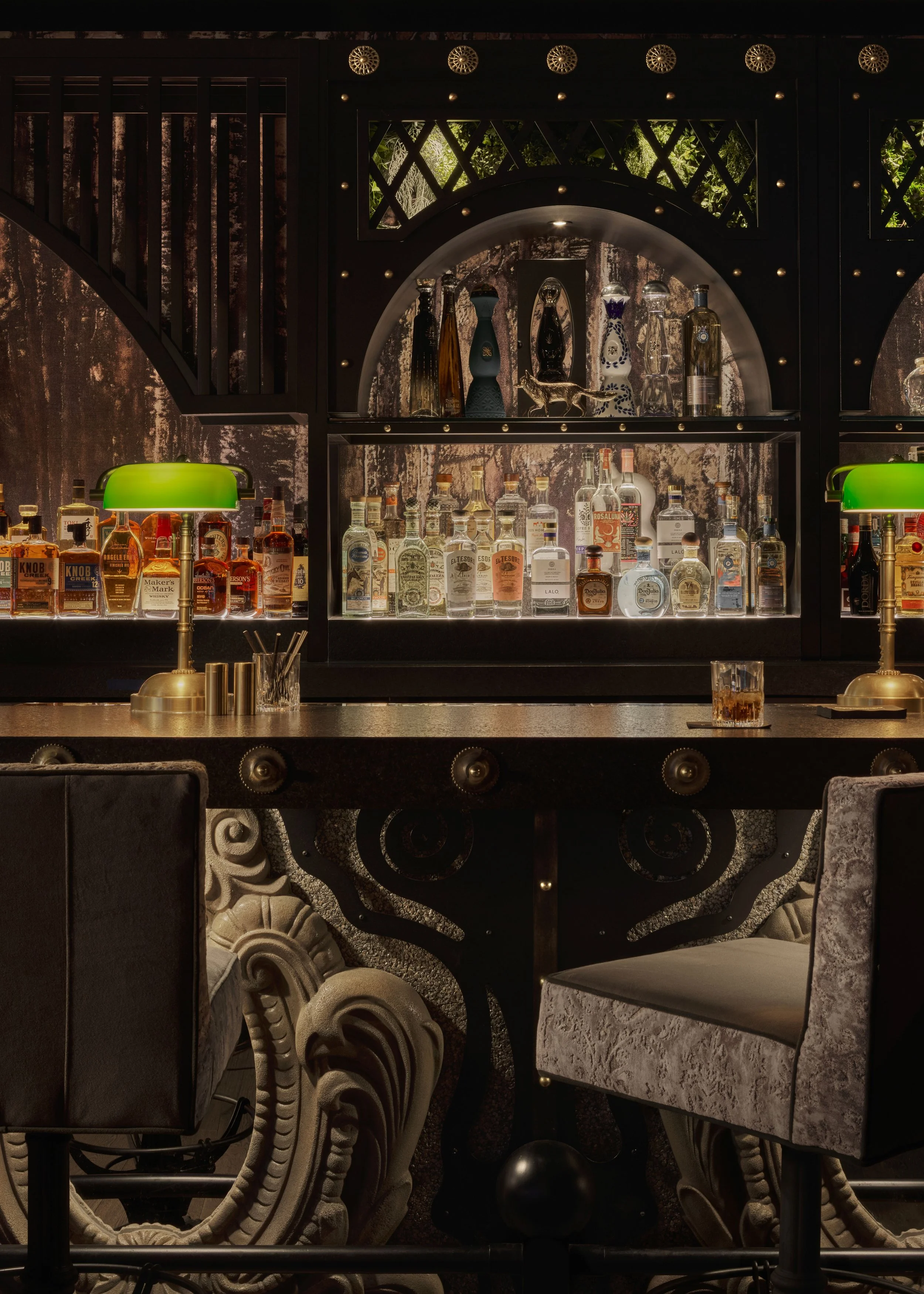 A bar with a black ornate counter, two green-shaded lamps, and shelves of various liquor bottles behind it. There are ornate chairs in the foreground.