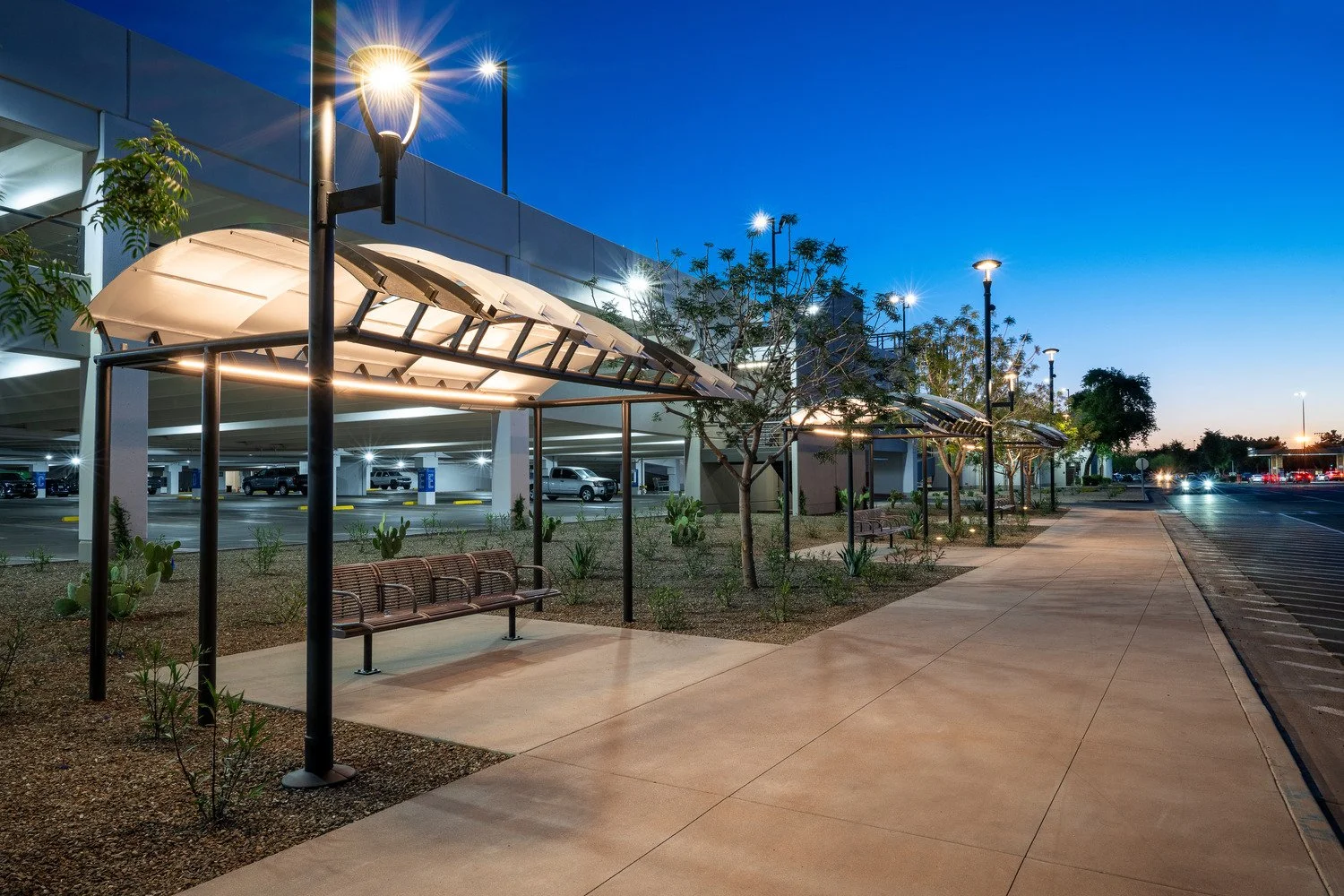 Empty sidewalk with benches and trees illuminated by streetlights outside a parking garage at dusk.
