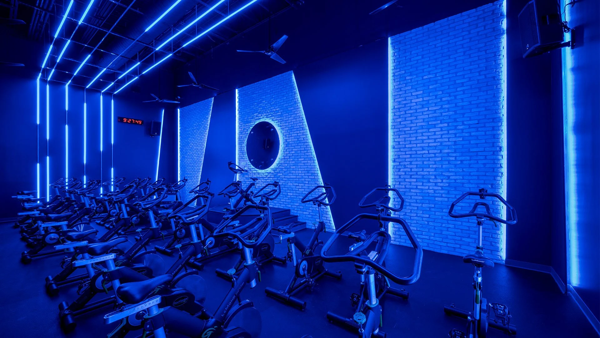 Indoor spinning bike gym illuminated with blue LED lights, brick wall accents, ceiling fans, and a digital clock, showing 9:27.