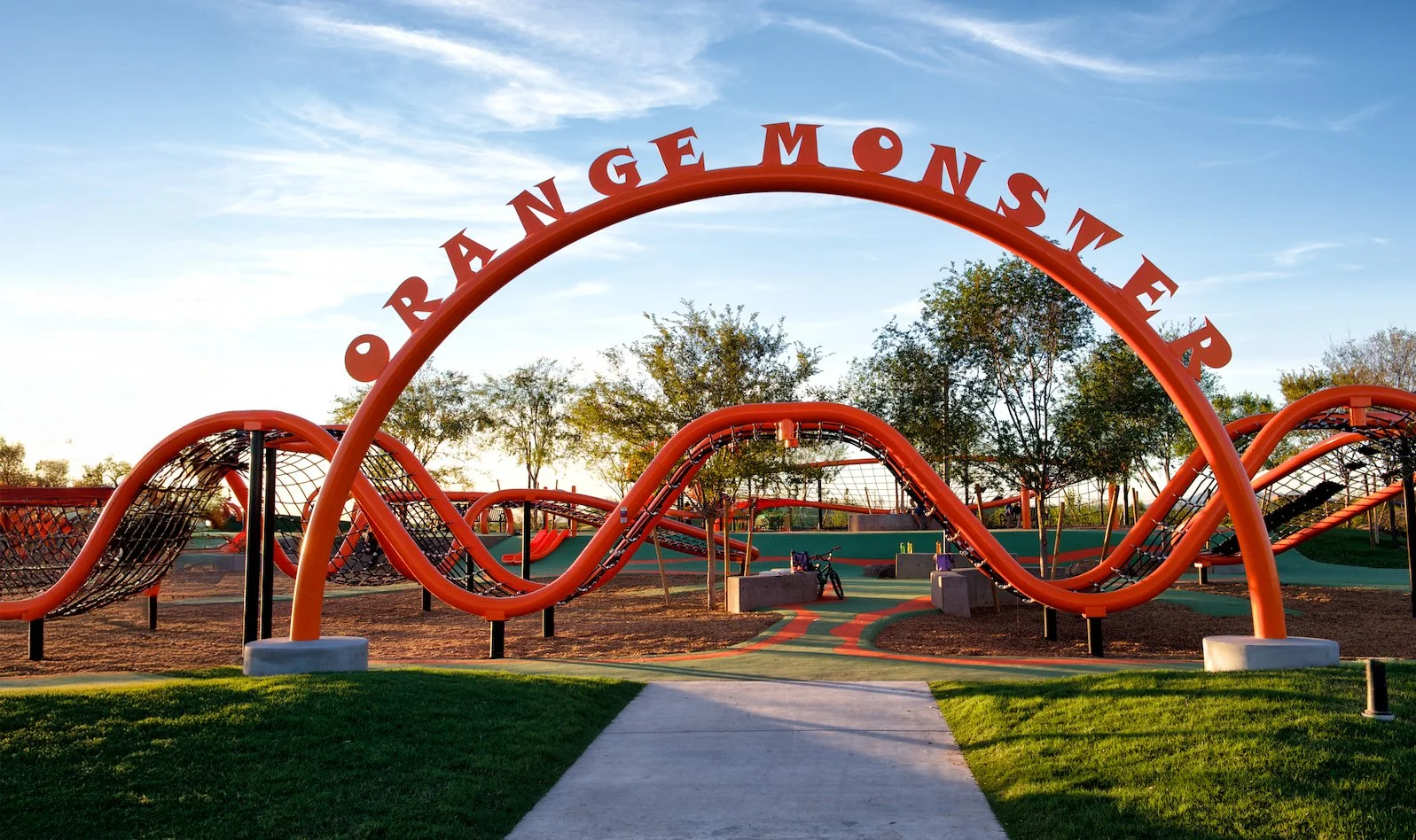 A playground with orange roller coaster-themed structures and a large arch that says 'Orange Monster' in a park, with trees and a blue sky in the background.