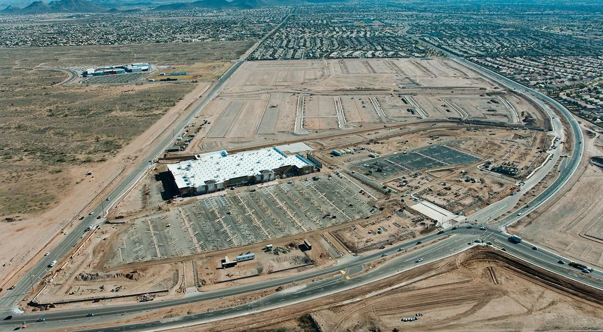 Aerial view of a large construction site with a mostly built shopping mall, parking lots, roads, and surrounding land, in an urban area.