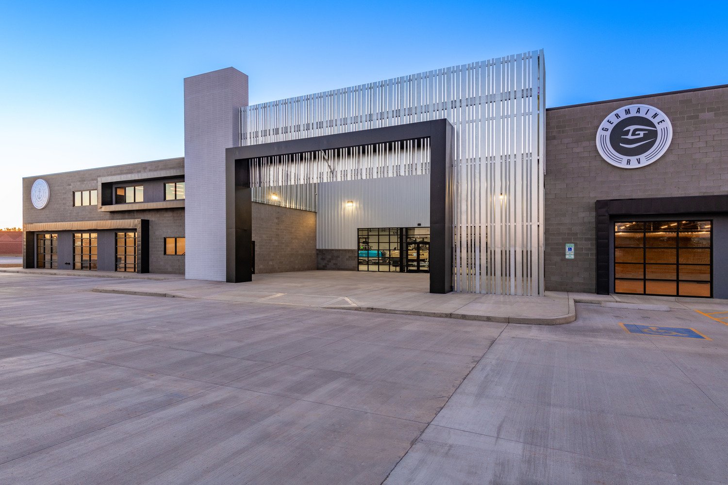 Front view of a modern commercial building with an open parking area, featuring black and gray exterior walls, large glass garage doors, and metal accents, with the Germaine RV logo on the upper right side.