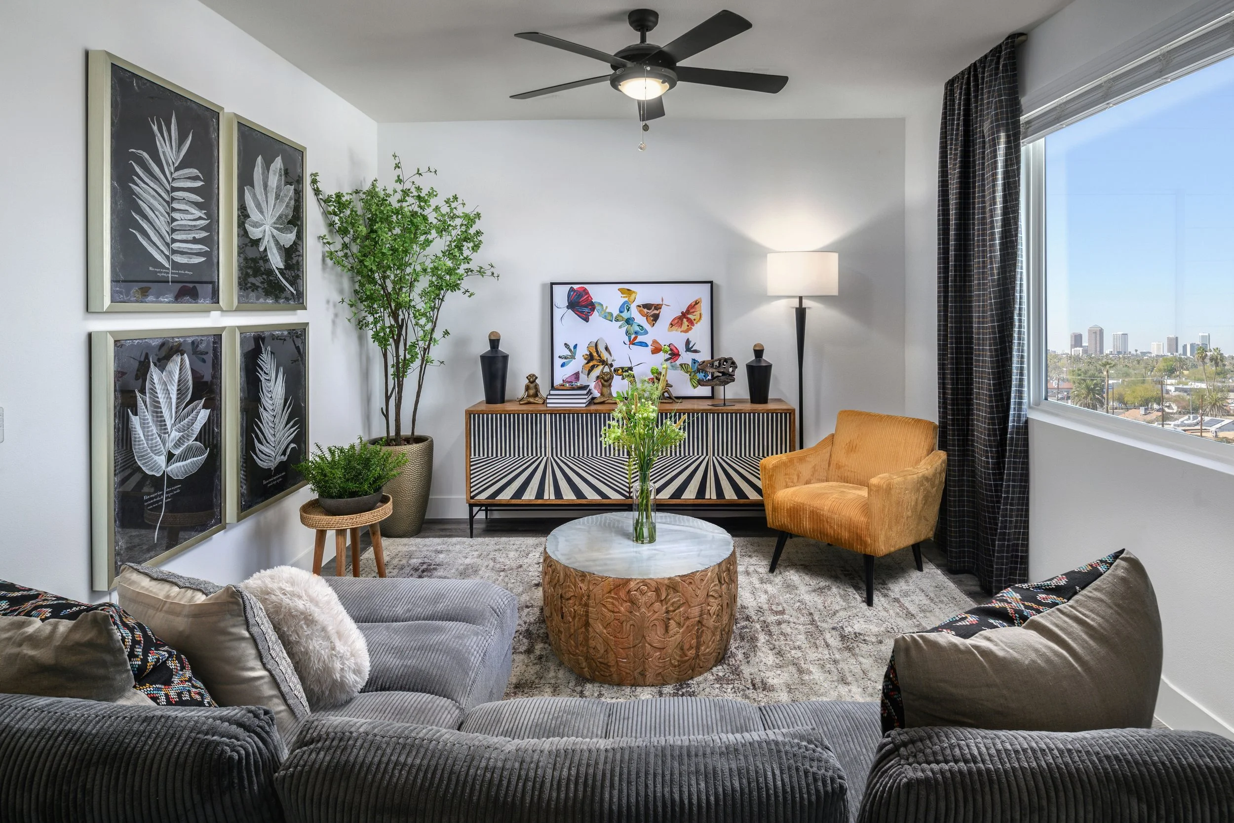 Living room with gray sectional sofa, yellow armchair, potted plants, framed butterfly artwork on wall, TV console with decorative vases, marble-topped coffee table with flower vase, window with black checkered curtains, city skyline view.