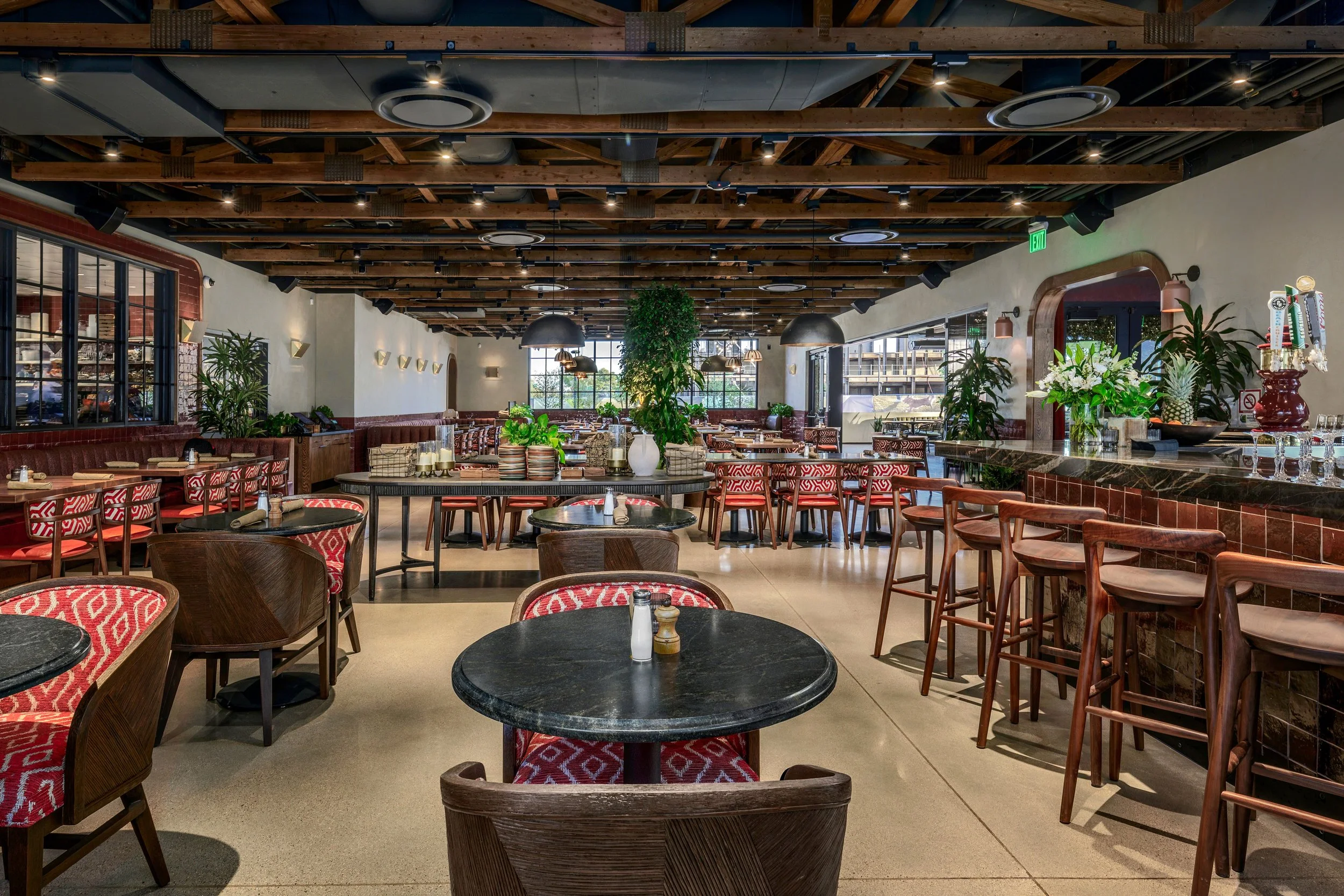Interior of a modern restaurant with wooden ceiling beams, black pendant lamps, red patterned chairs, black tables, and lush green plants, with large windows providing natural light.