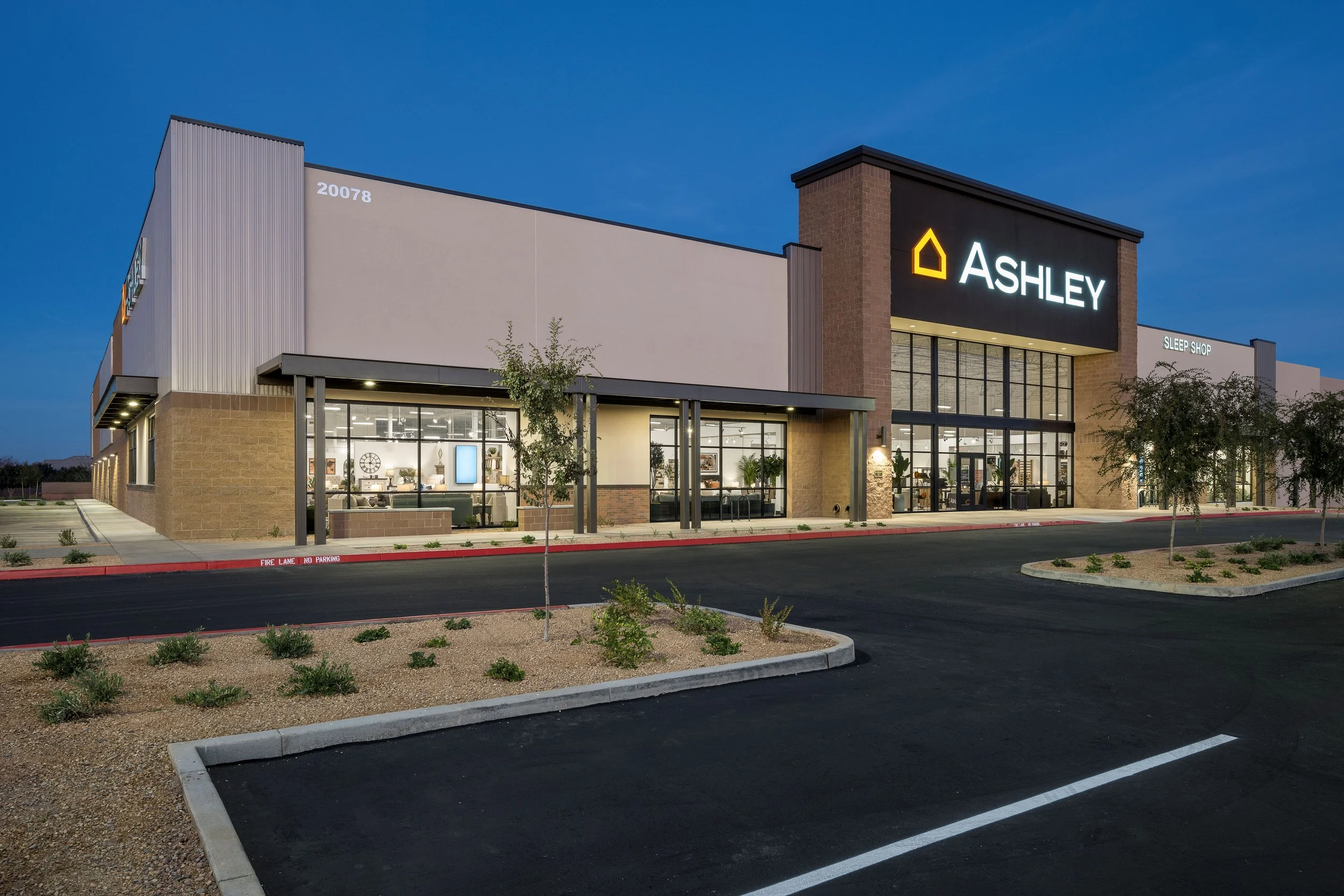 Exterior of a shopping center at dusk with a sign reading "Ashley" and a smaller sign "Sleep Shop." The parking lot is empty, and there are a few young trees and plants around the area.