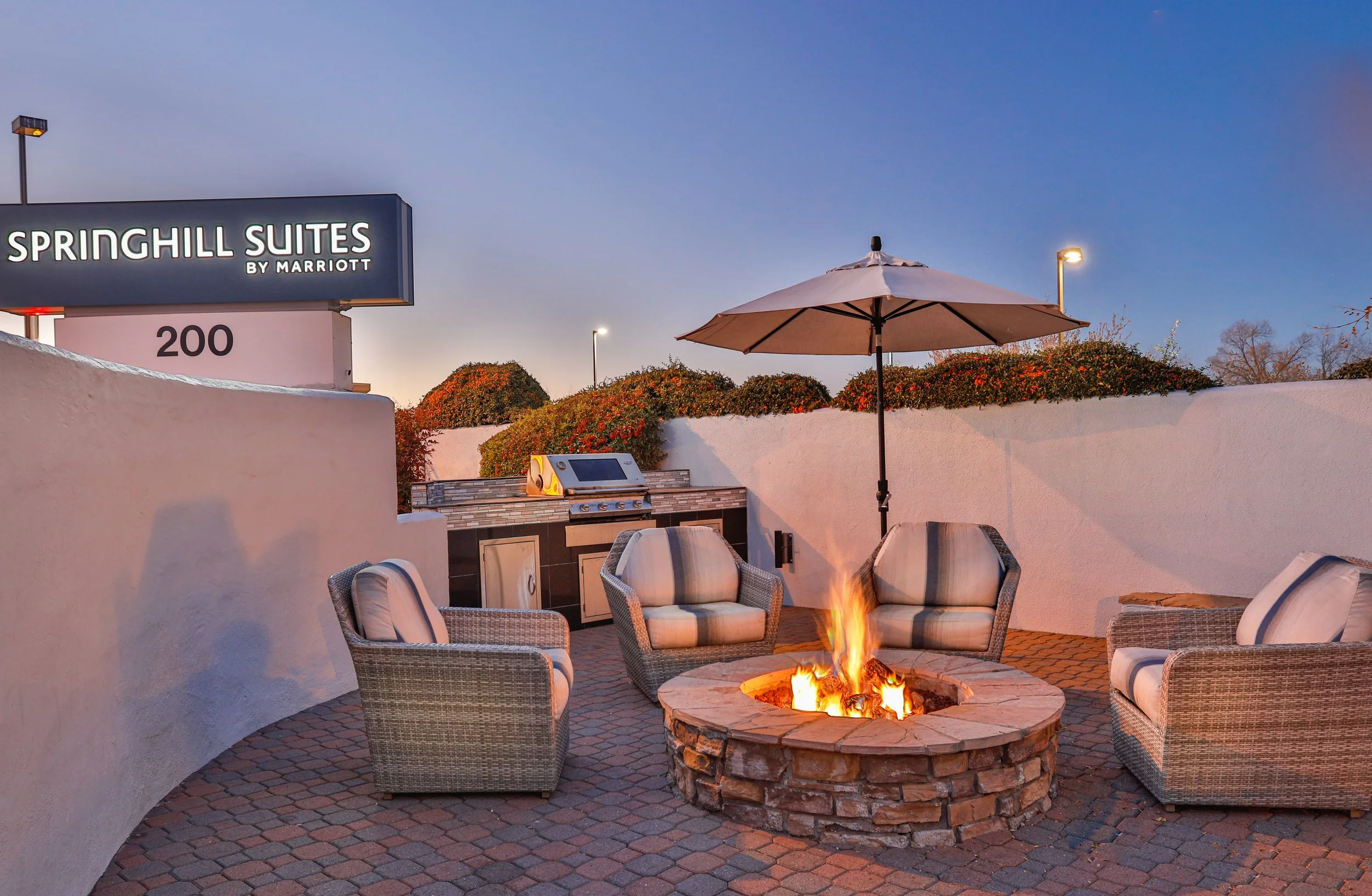 Outdoor patio area with four cushioned chairs around a fire pit, a large umbrella, a built-in grill, and a sign for Springhill Suites by Marriott in the background.