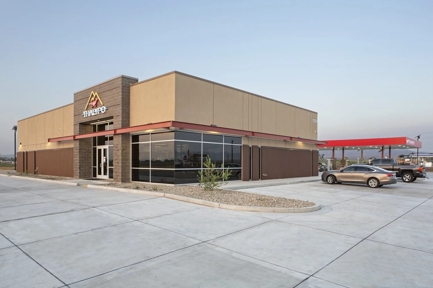 Wide view of a modern fast-food restaurant, ThaiExpress, featuring a beige and brown exterior with large glass windows, a red canopy over the drive-thru, and a parking lot with a few cars.