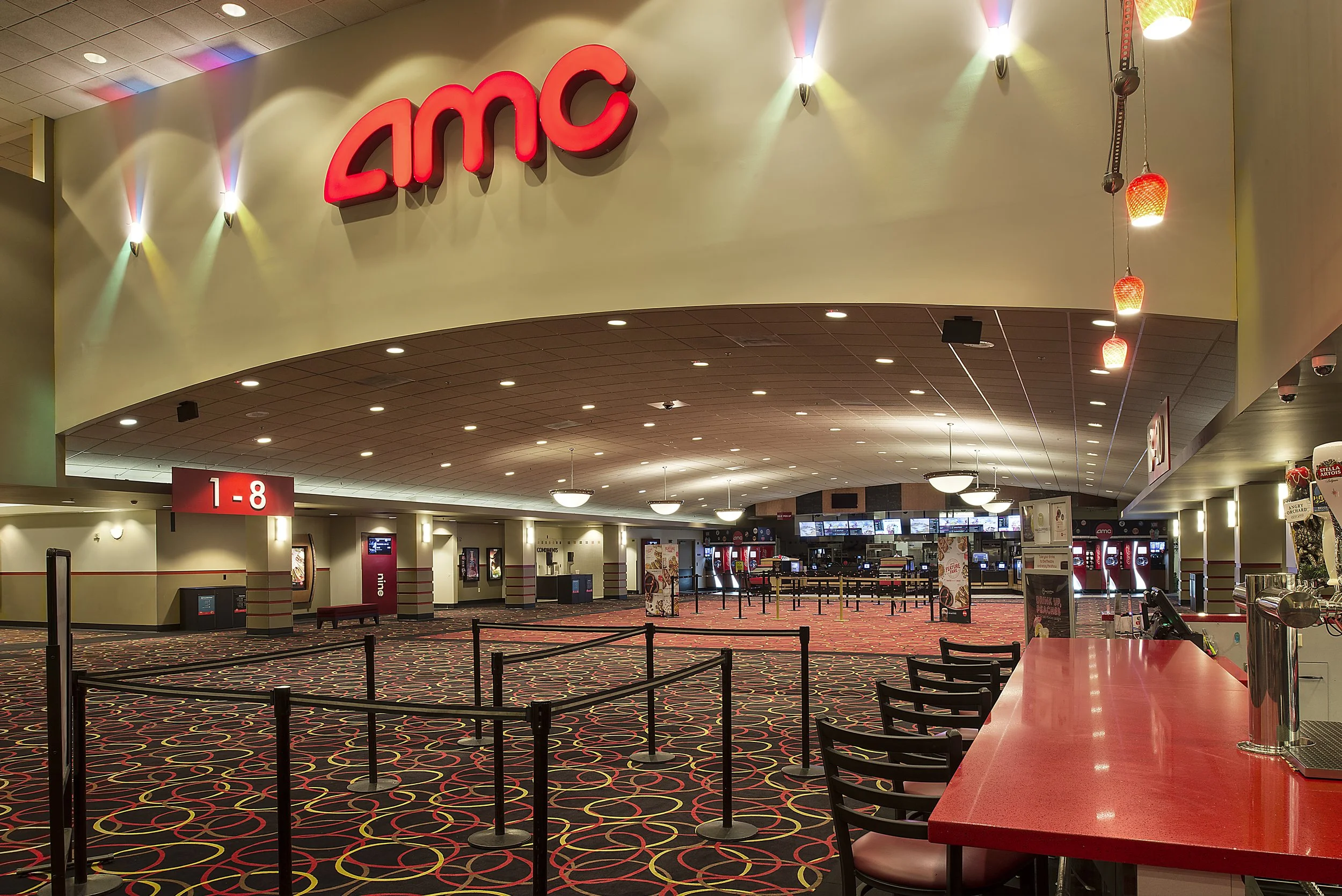 Interior of an AMC movie theater lobby with a red counter, chairs, and digital screens showing movie listings, with a large AMC sign overhead.