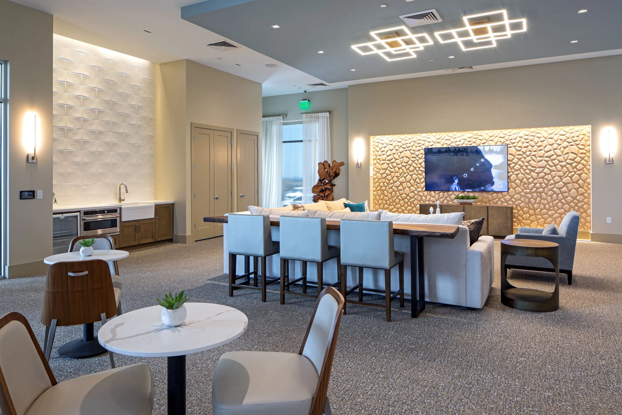 Modern living room with a large beige sectional sofa, a flat-screen TV mounted on a patterned wall, and a bar-height table with five chairs. The room has a kitchenette with a sink, microwave, and cabinets, as well as small round tables with potted su