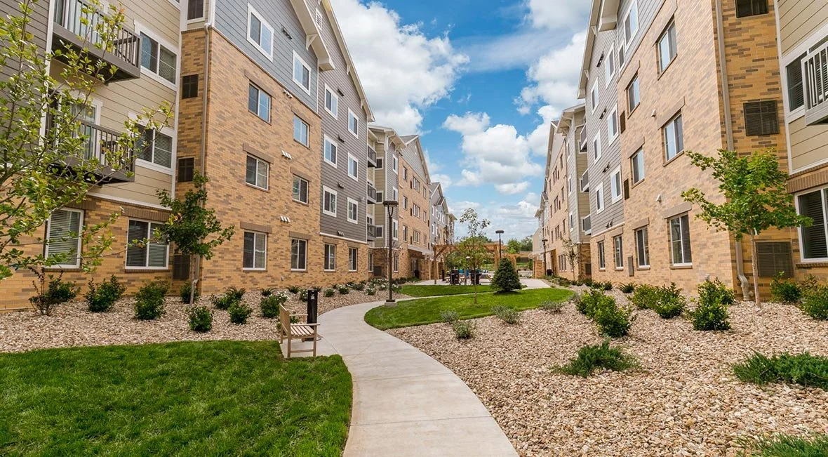 View of a residential apartment complex courtyard with a curved concrete pathway, green grass, small bushes, trees, and benches, with multi-story buildings on either side and a partly cloudy sky overhead.