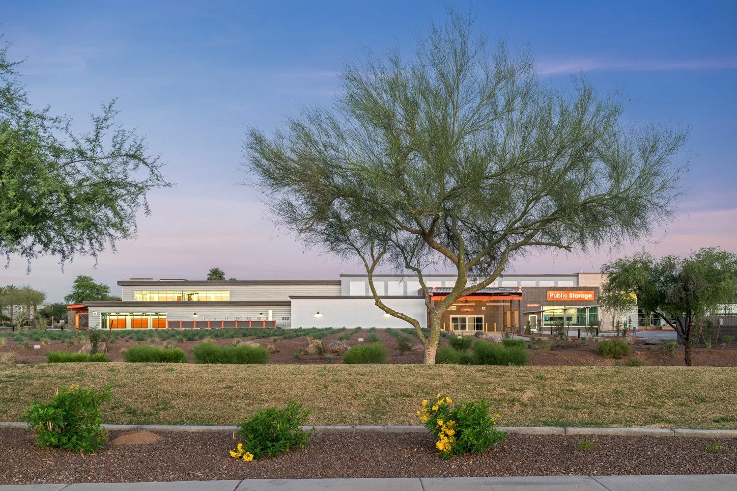 Exterior view of a modern public storage facility with a large tree and landscaped bushes in the foreground, and a sign reading ‘Public Storage’ on the building, during dusk.