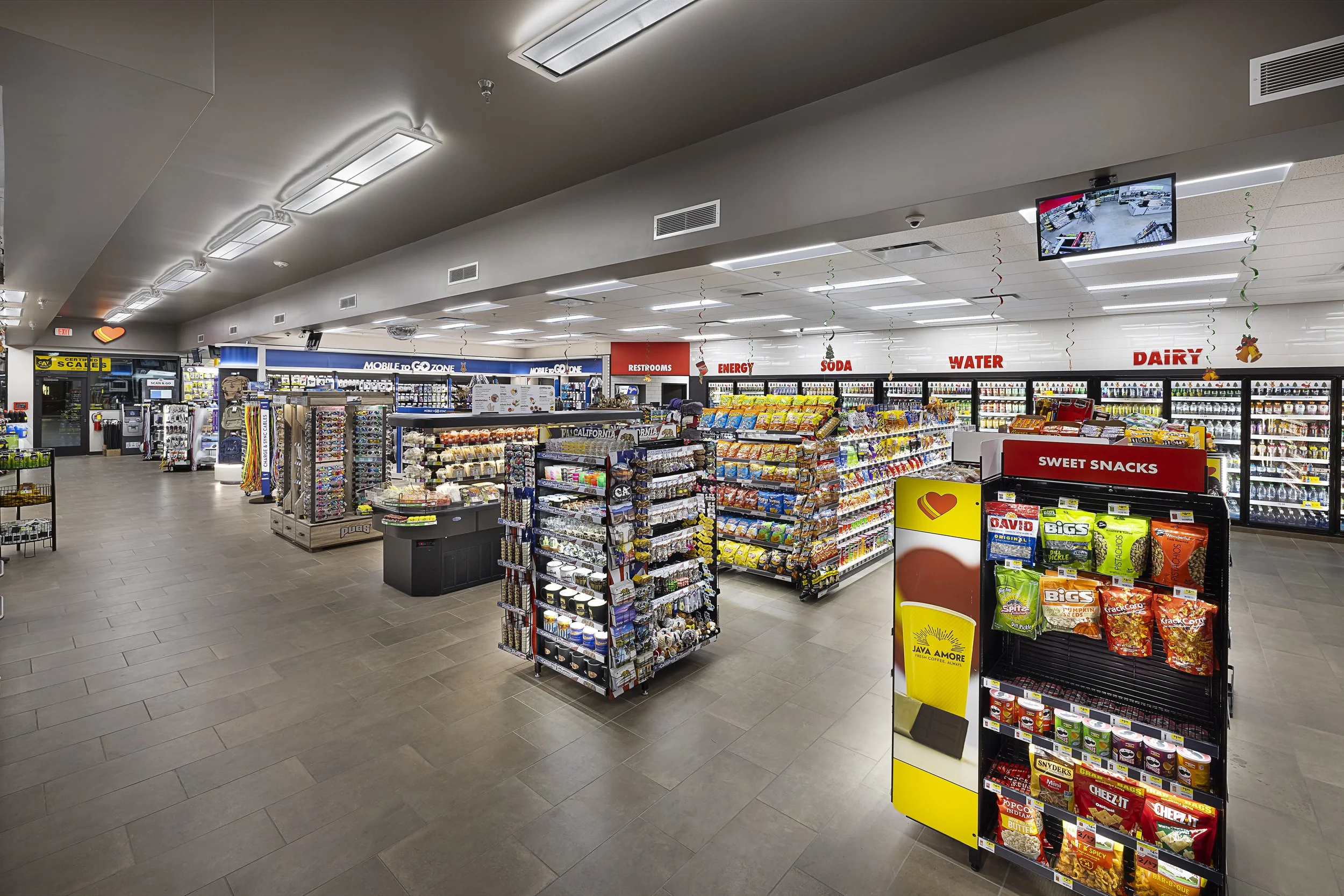 Interior of a convenience store with shelves stocked with snacks, beverages, and other items. Signs indicate sections for energy drinks, soda, water, dairy, and sweet snacks, along with a digital monitor on the ceiling.