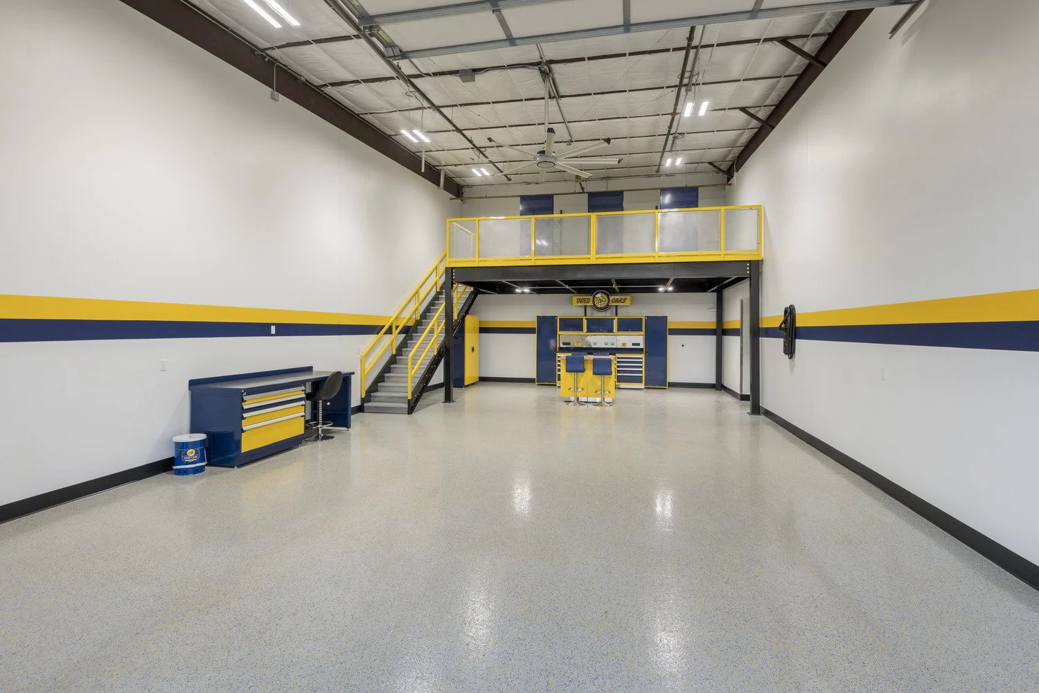 Empty workshop or garage with a yellow and blue color scheme, featuring a workbench, tool storage, and a loft area accessed by a staircase.