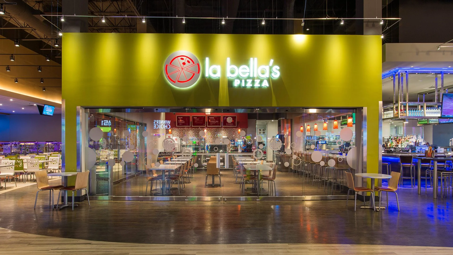 Interior view of La Bella's Pizza restaurant with a bright green exterior sign and seating area visible through glass walls, inside a shopping mall or entertainment complex.