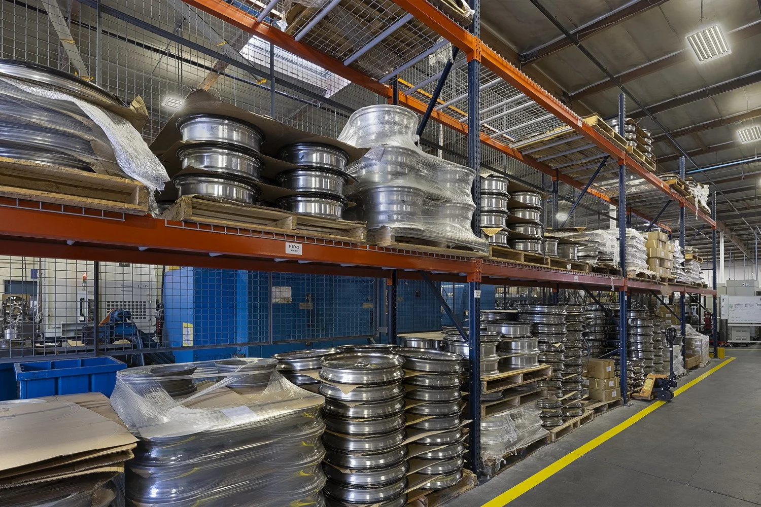 Industrial warehouse storage shelves filled with metal machine parts, some wrapped in plastic, with a forklift on the right side, concrete floor, and ceiling with lights.