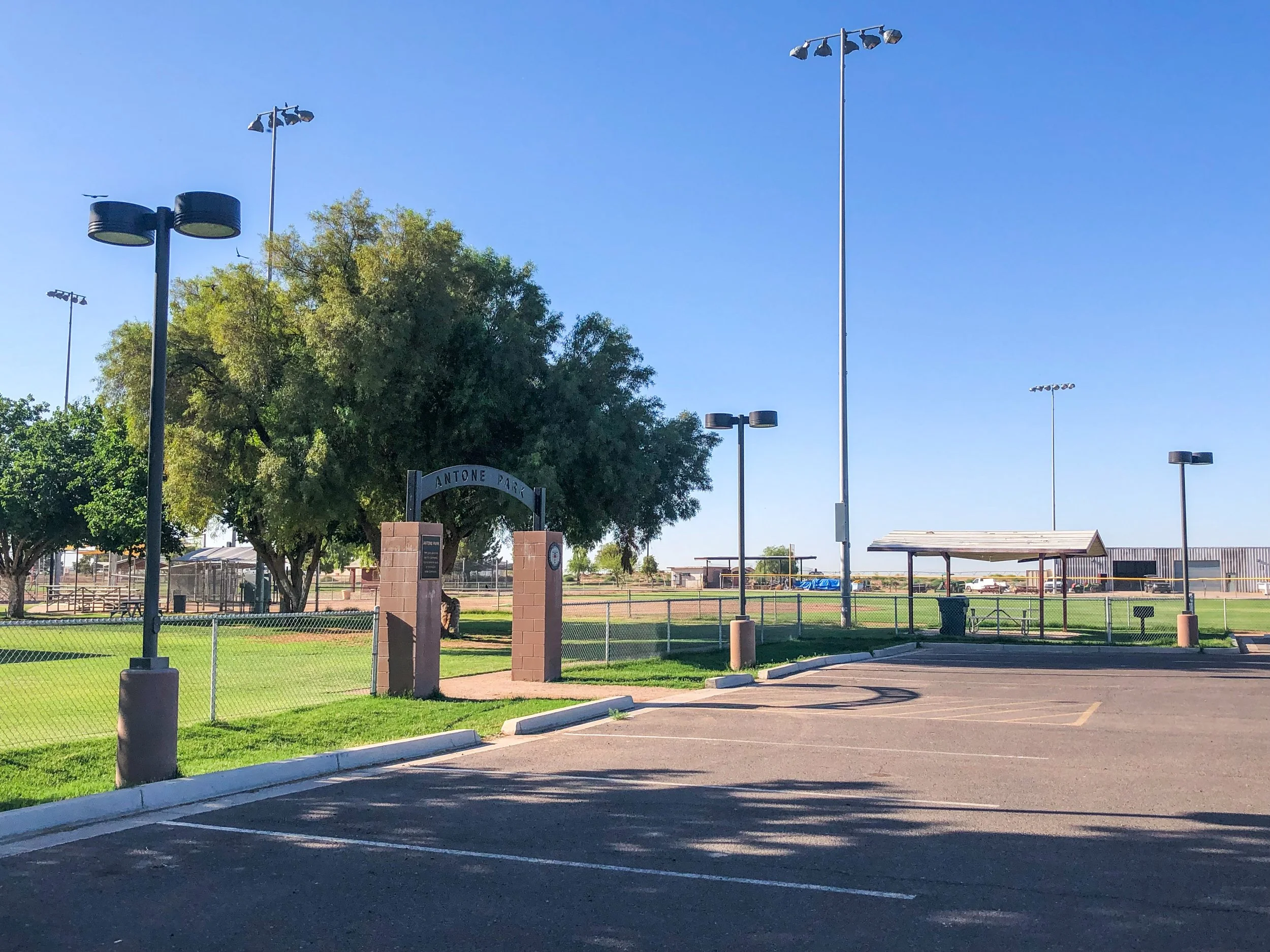 Empty parking lot near a park with a sign that reads 'Antone Park,' trees, and sports fields in the background under a clear blue sky.