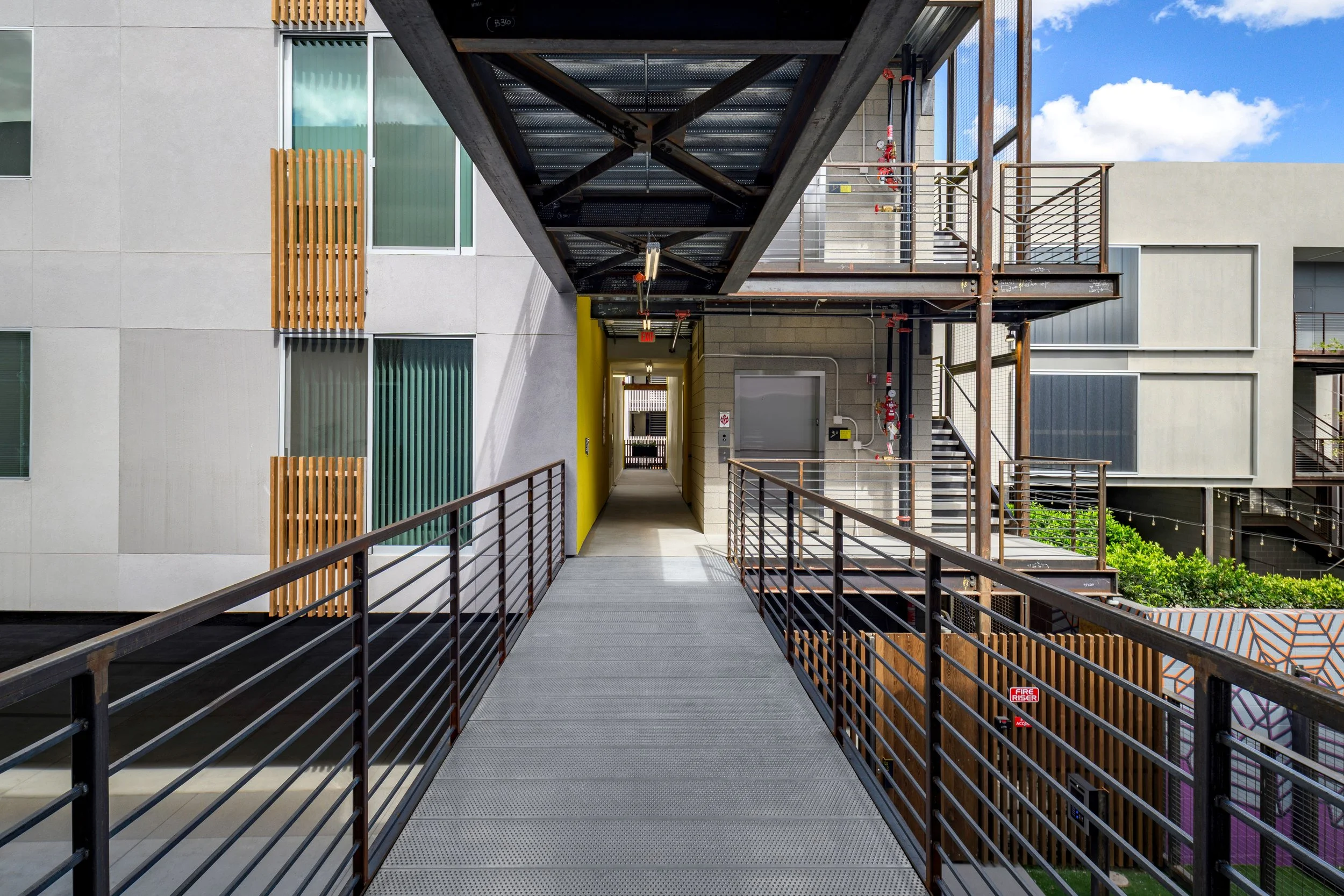 Photo of a modern apartment balcony walkway with metal railings, leading to stairs and a doorway, with neighboring building walls and a blue sky with clouds in the background.