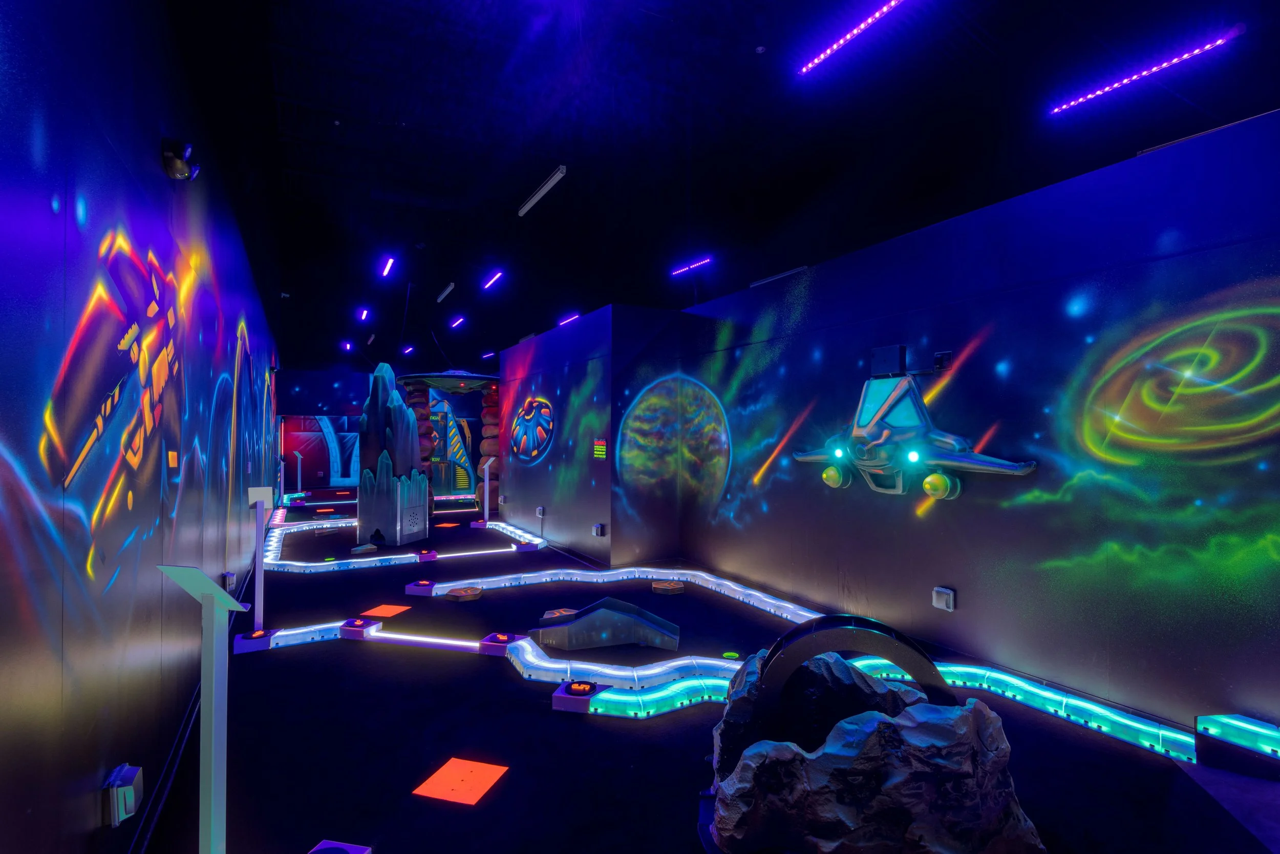 Indoor mini golf course with neon space-themed wall murals, glowing obstacles, and a spaceship decoration.