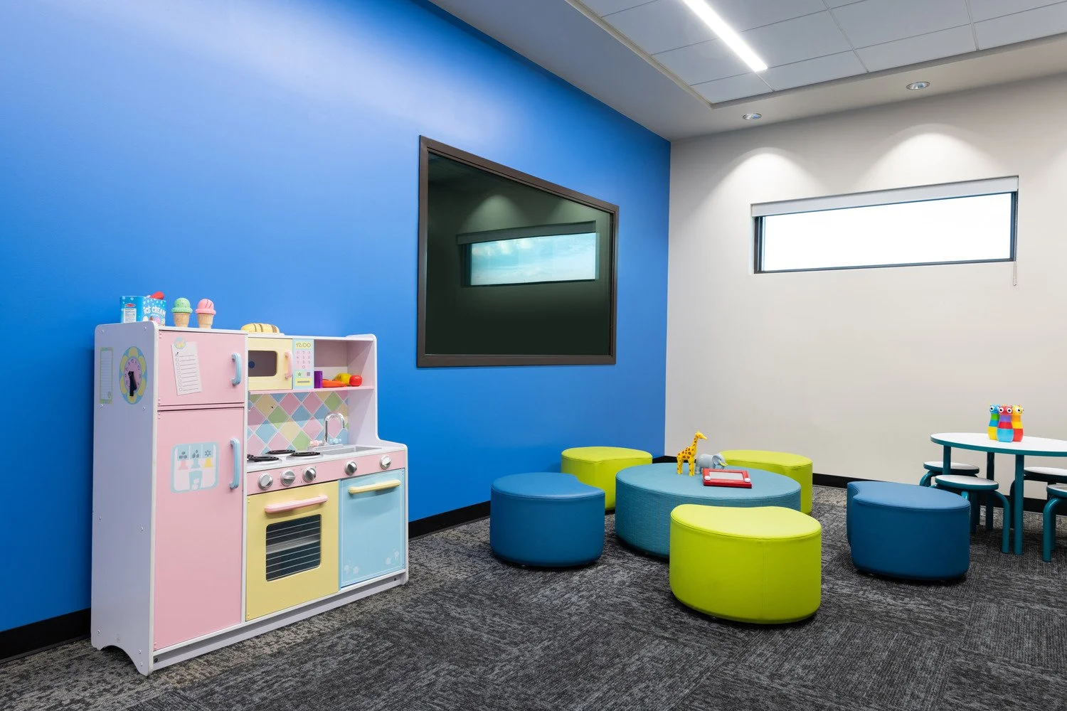 Colorful children's playroom with a pastel pink, yellow, and blue toy kitchen set, green and blue ottomans, a small round table with stools, toy animals including a giraffe, a zebra, and an owl, and bright windows with natural light.