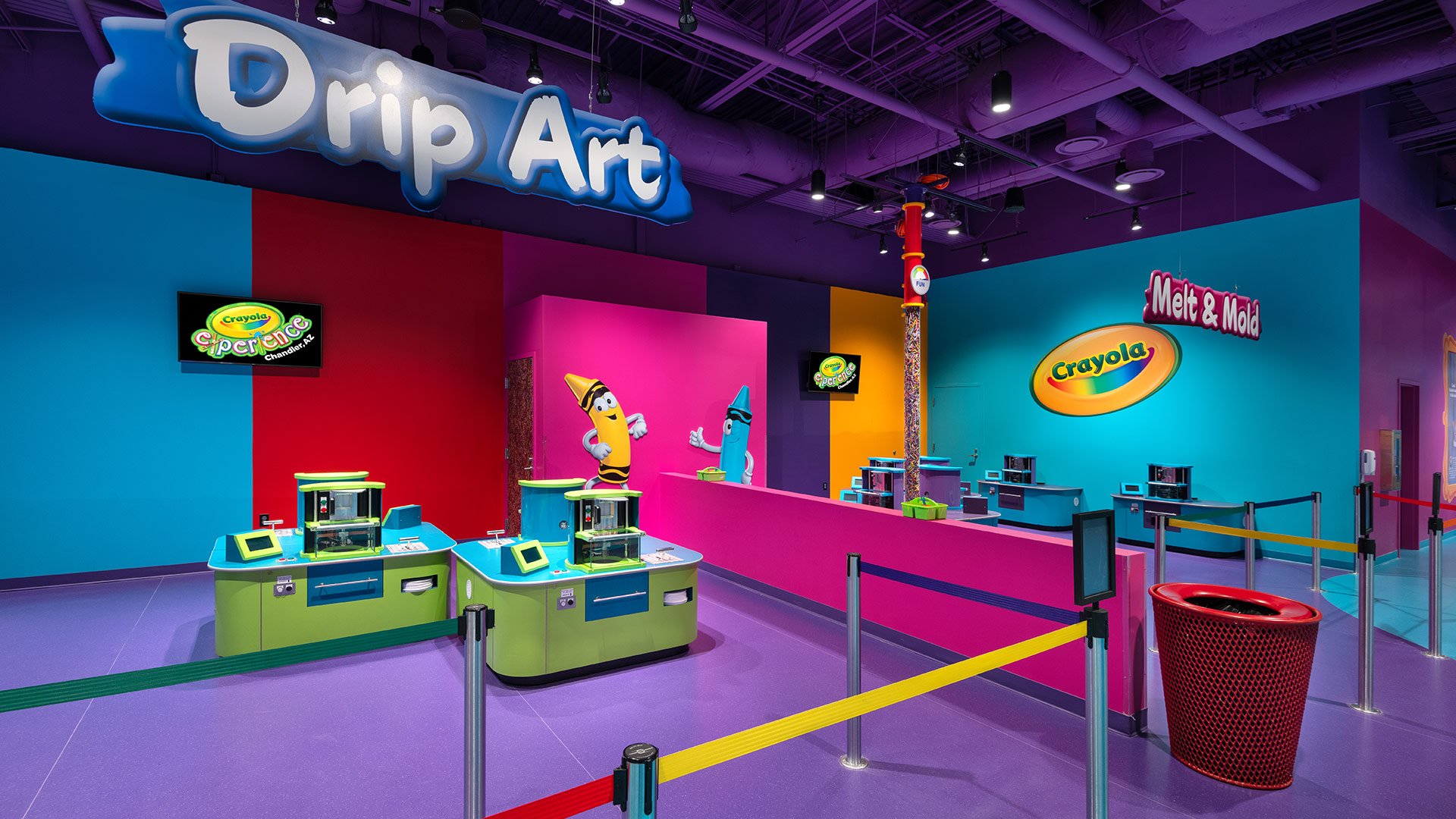 Colorful indoor Crayola experience area with a large sign that says 'Drop Art'. There are display screens, themed decorations of cartoon crayon characters, and a rope barrier sectioning off part of the space, with a red trash can nearby.