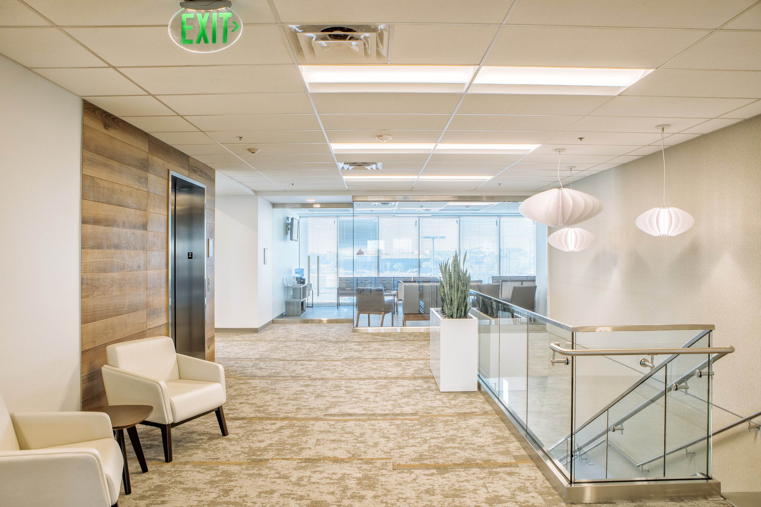 Empty modern lobby area with seating, an elevator with wood paneling, a large window, a potted cactus plant, hanging white pendant lights, and an emergency exit sign.