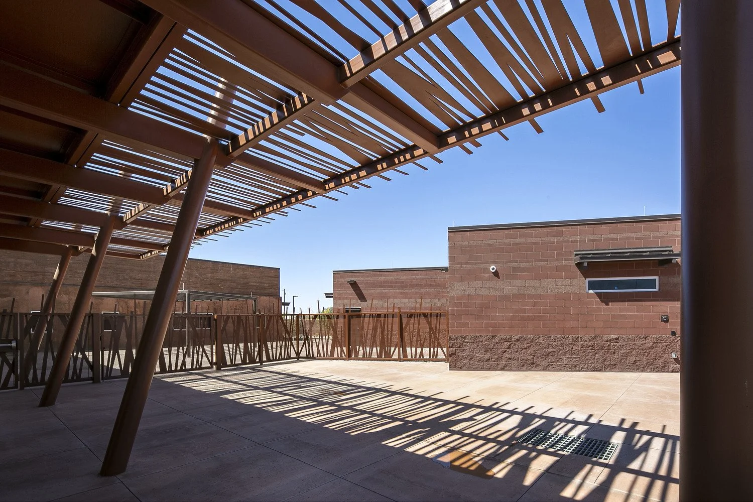 An outdoor patio area with wooden slatted shade structure casting shadows on the ground, and a brick building in the background under a clear blue sky.