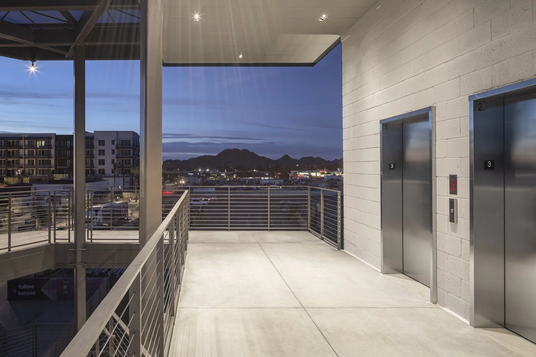 Empty outdoor balcony area with two elevators on a brick wall and a view of a cityscape with mountains in the distance at dusk.