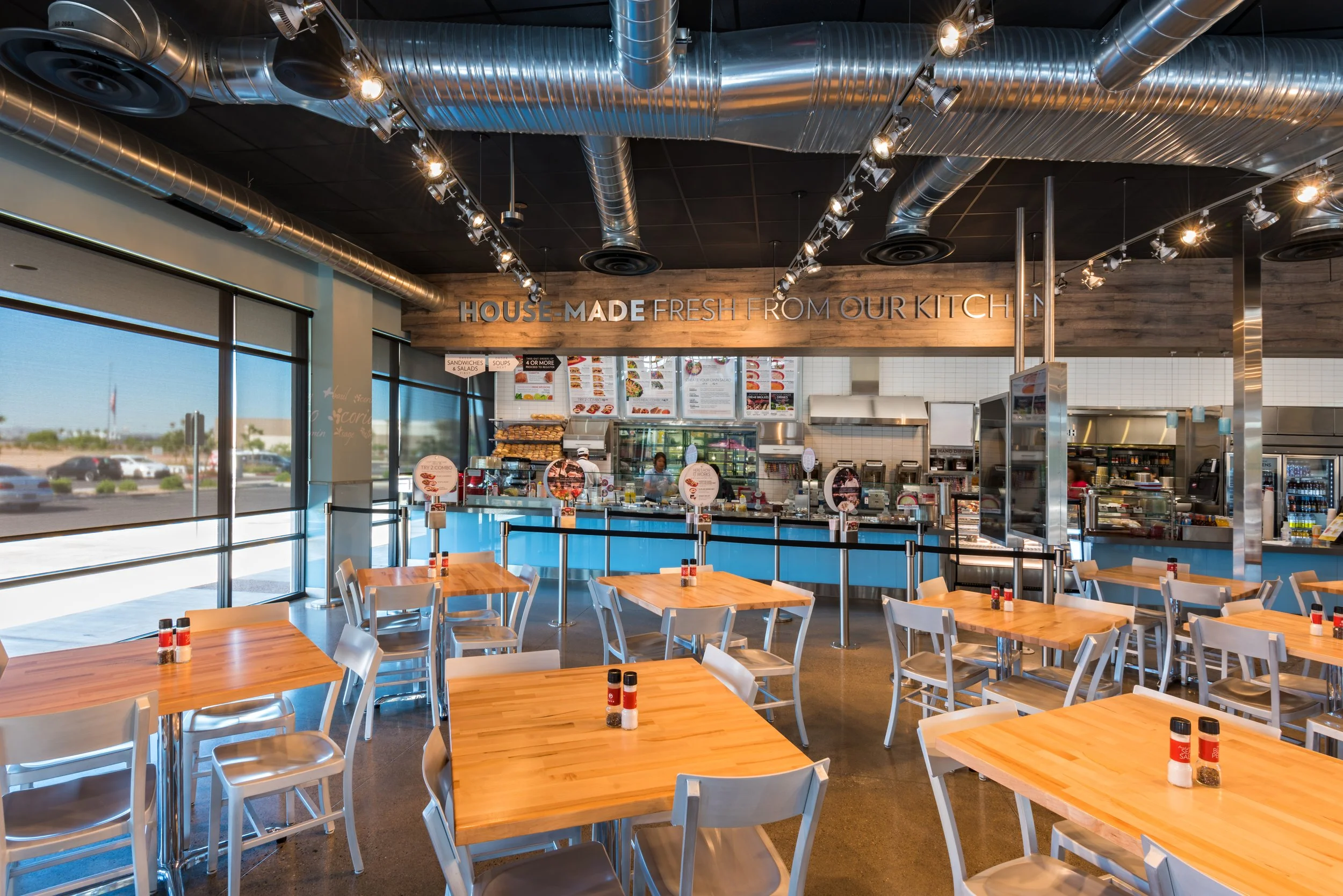 Interior of a modern fast-food restaurant with wooden tables, metal chairs, large windows, and exposed ductwork ceiling; the counter area has menu boards and a sign that reads 'HOUSE-MADE FRESH FROM OUR KITCHEN'.