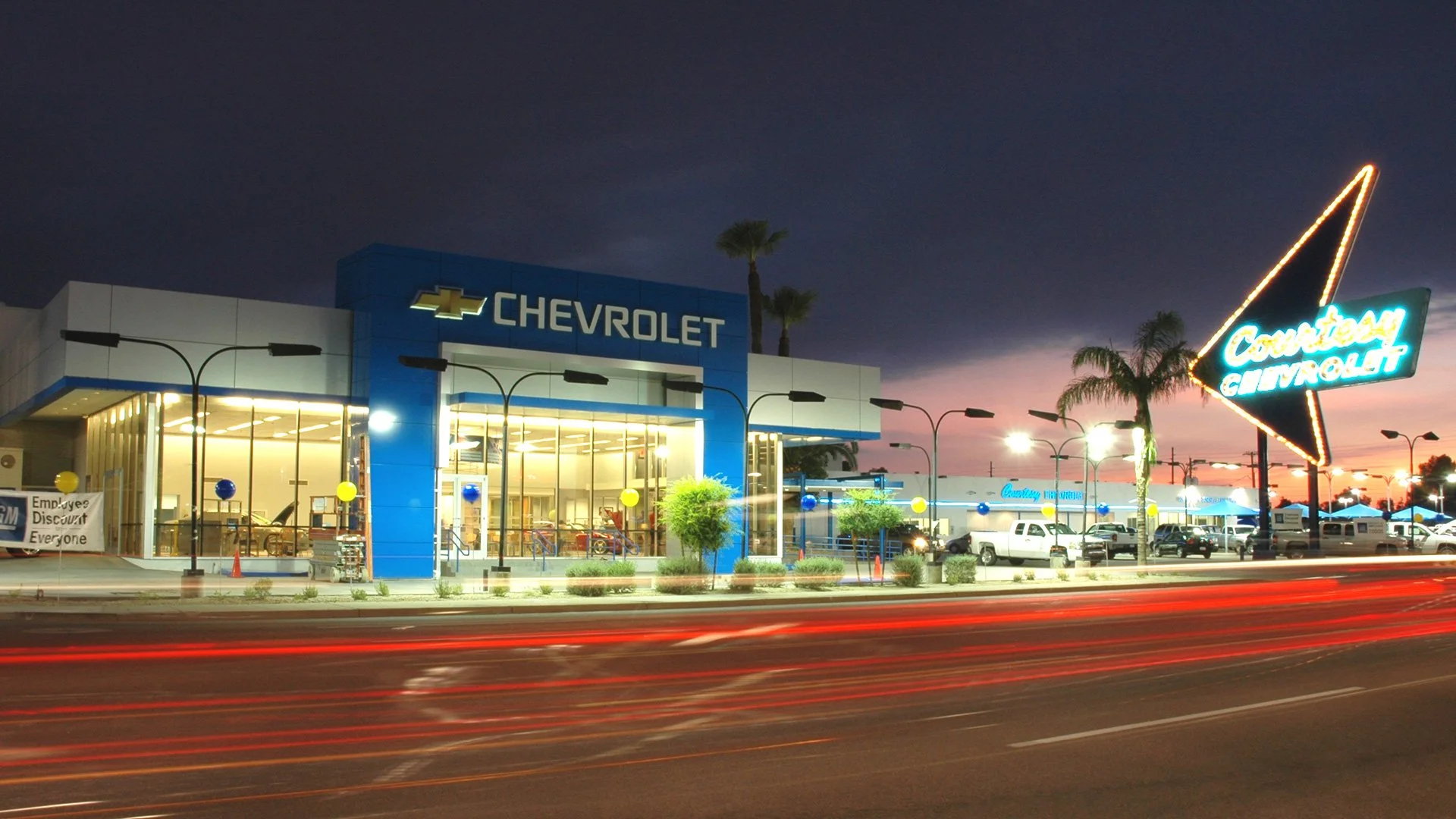 Exterior of a Chevrolet dealership at dusk with a large Chevrolet sign, bright indoor lighting, and streaks of red light from passing cars on the street.