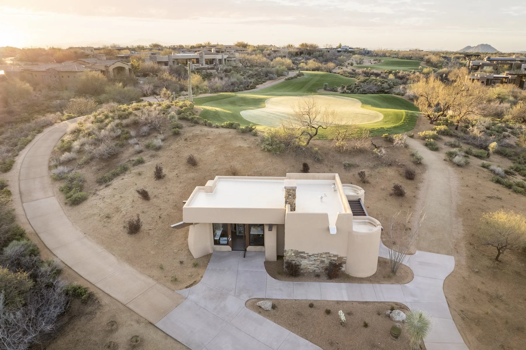 Aerial view of a modern house with an outdoor area, surrounded by desert landscaping, near a golf course with a sand trap and green grass, in a desert landscape.