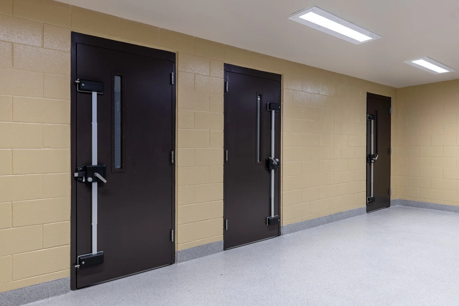 Three black emergency exit doors with crash bars, installed on a beige brick wall in a hallway with gray carpeted flooring and fluorescent ceiling lights.