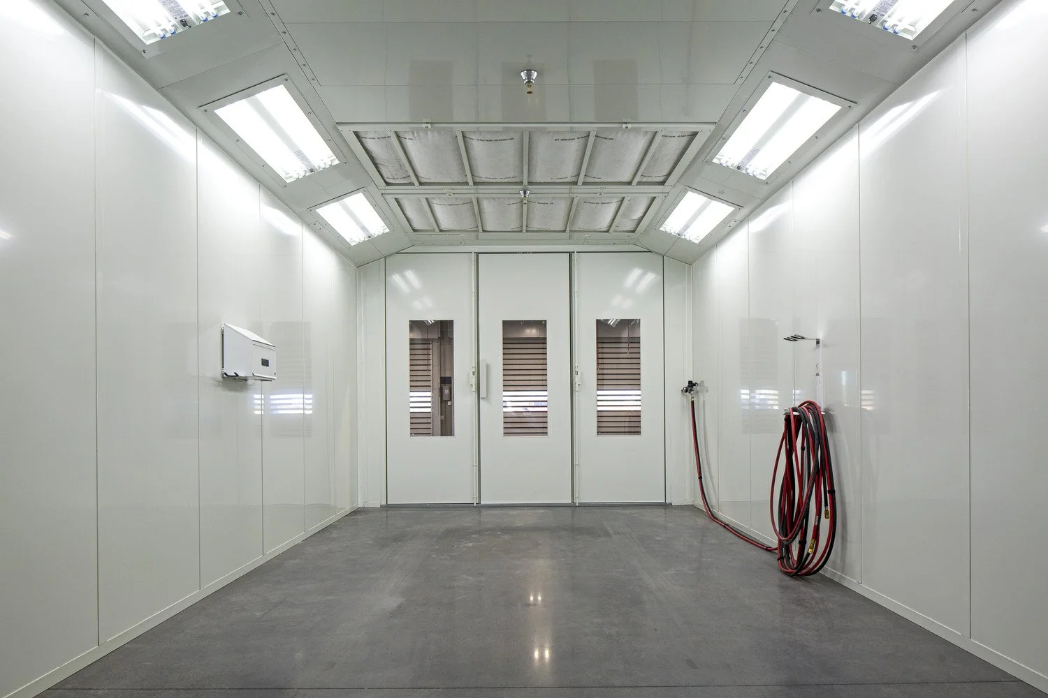 Empty spray paint booth with white walls, grey floor, and ventilation system on the ceiling, with red and black hoses on the right wall.