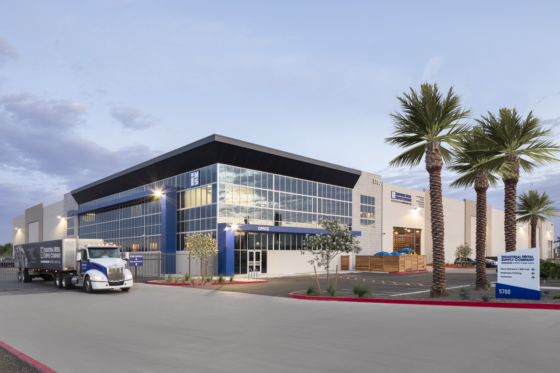 Modern industrial building with large glass windows, palm trees in the parking lot, and a semi-truck parked outside. There are signs indicating the location of offices and delivery areas.