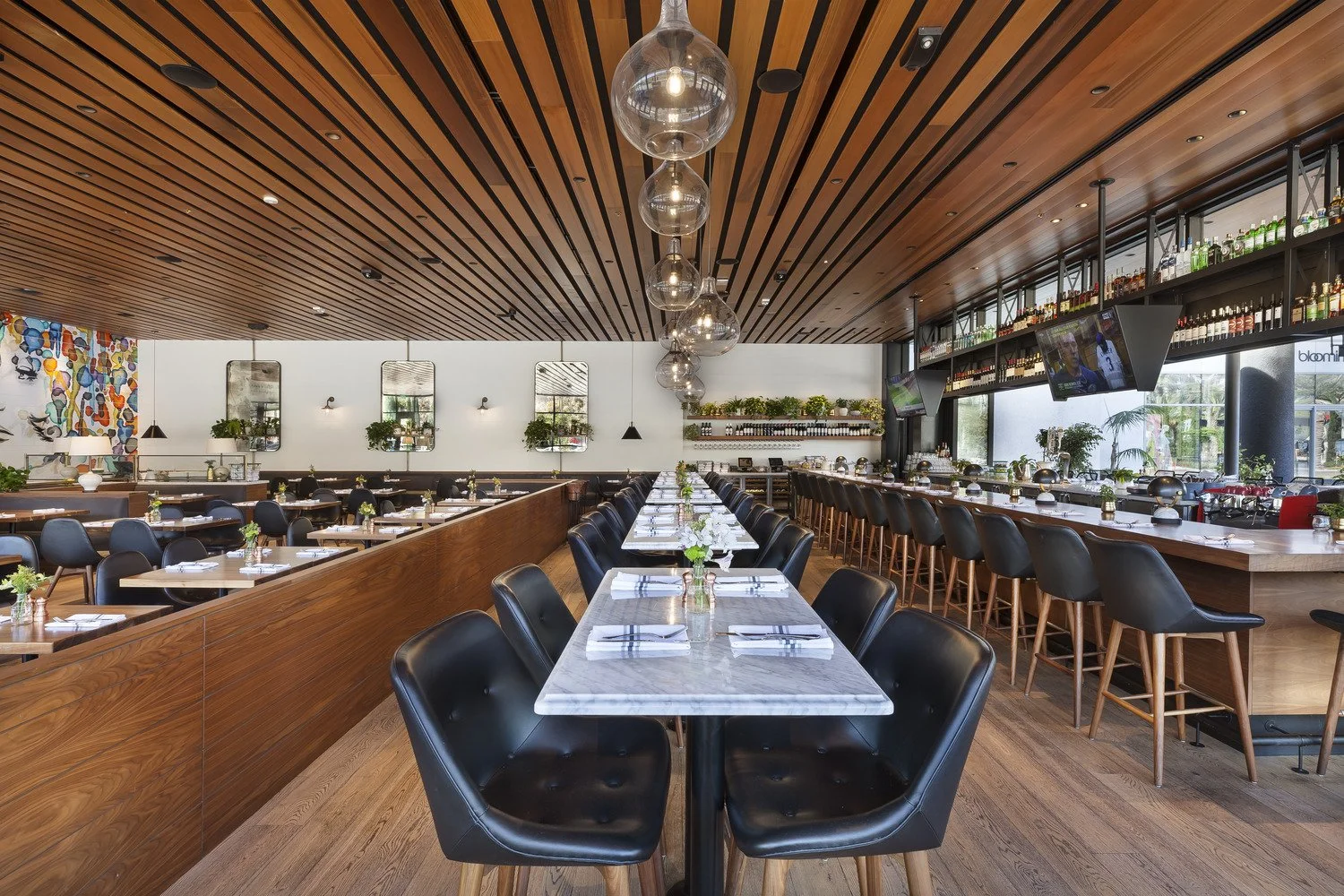 A modern restaurant interior with wooden ceiling, black chairs, and white marble tables set with napkins and cutlery, decorated with small green plants, with a bar area and flat screen TVs on the wall.