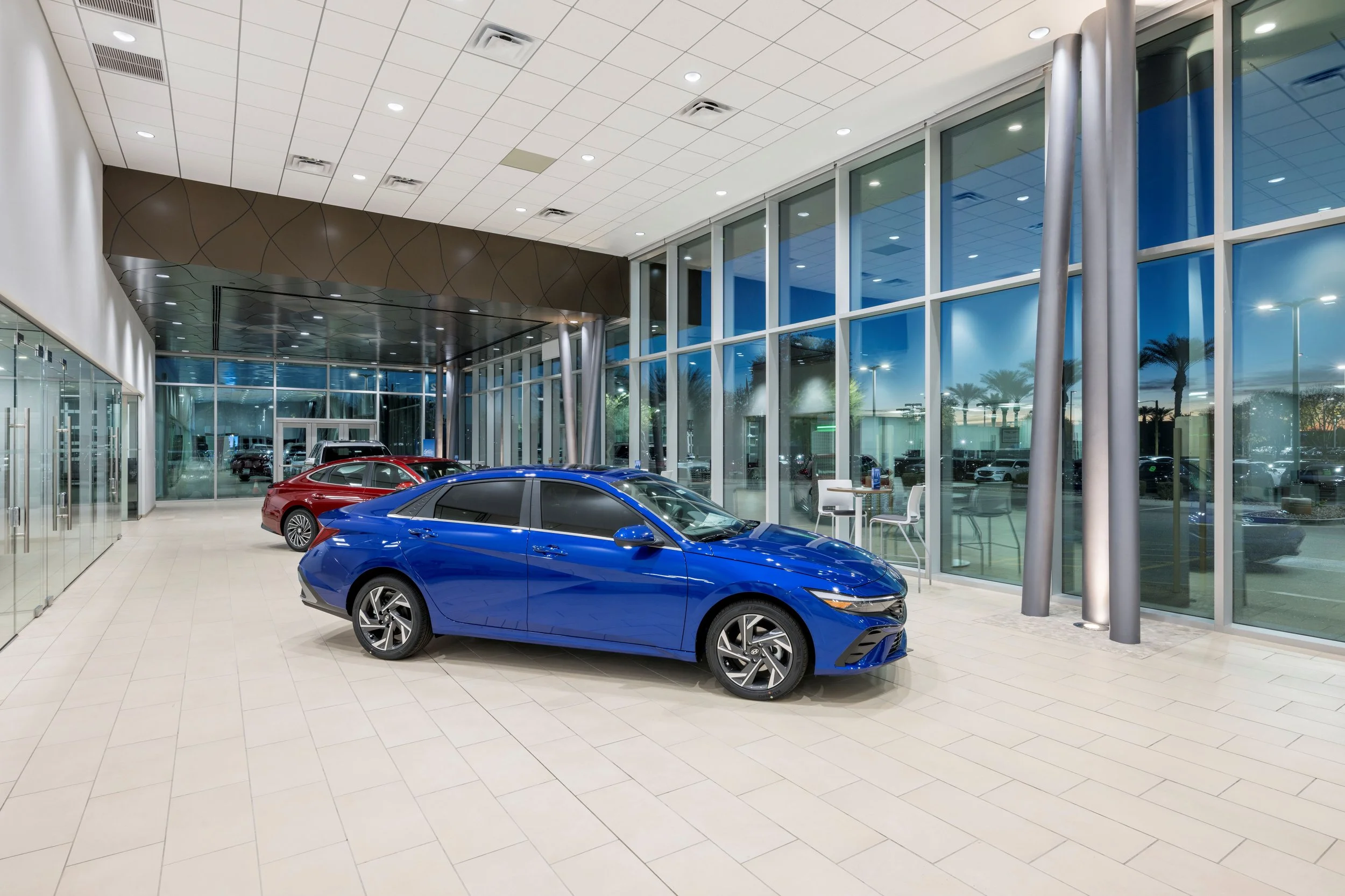 Interior of a modern car dealership showroom with glass walls, a blue sedan in the foreground, and a red sedan behind it. Large windows reveal palm trees and a parking lot outside at dusk.